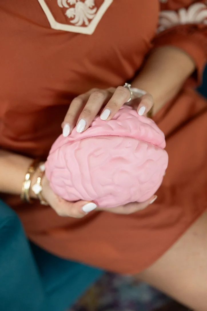A person holding a pink brain model with both hands, wearing white nail polish and gold jewelry, with a rust-colored shirt featuring a white crest.