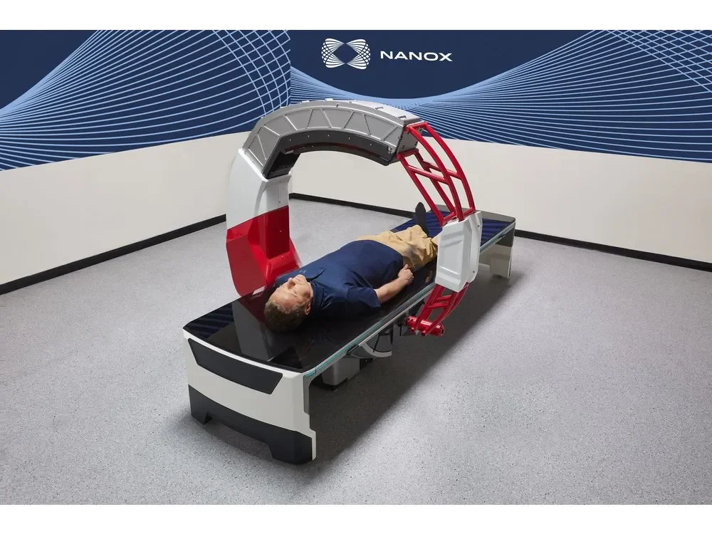 A man lying on a robot-assisted medical imaging table with a large circular scanning device positioned above him, in a testing room with a blue wave-patterned wall and the logo 'NANOX'.