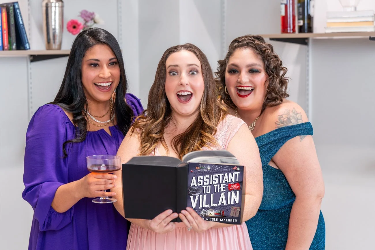 Three women reading the book Assistant to the Villain by Hannah Nicole Maehrer and enjoying cocktails
