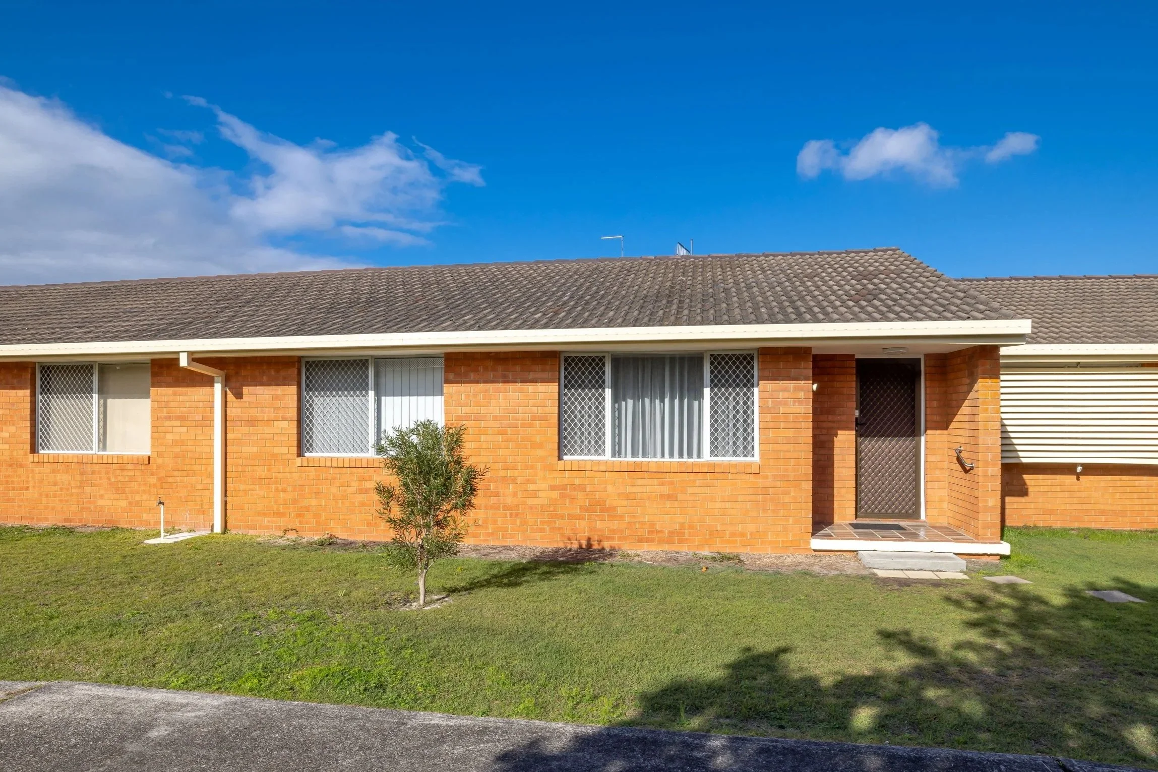 Front view of a single-story brick house with three windows, a small staircase, and a tree in front, under a blue sky with some clouds.