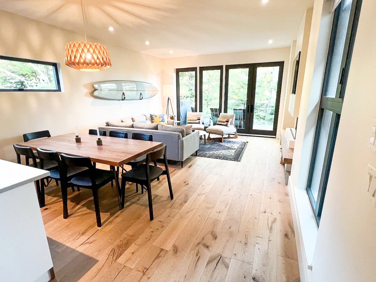 Open-plan living room and dining area featuring a wooden dining table with black chairs, a beige couch, modern chairs, and a surfboard wall decoration. The room has large windows and wooden flooring, creating a bright and airy atmosphere.