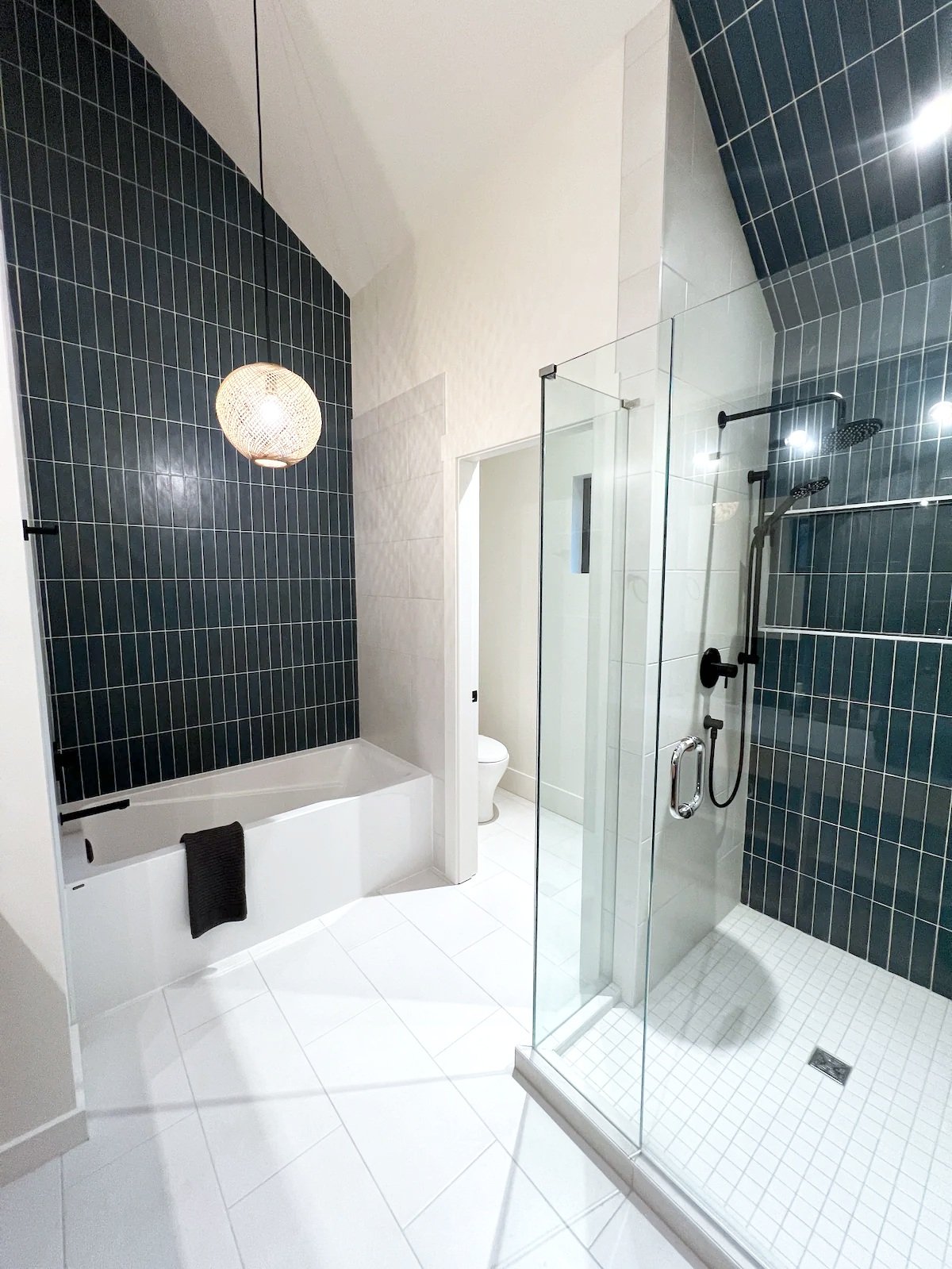 Modern bathroom with white bathtub, glass shower enclosure, dark blue tile walls, white tile floor, and round pendant light.