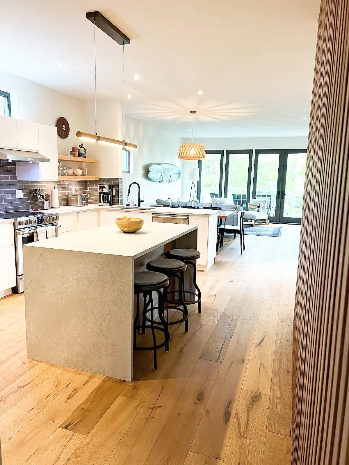 Modern kitchen and dining area with hardwood floors, a kitchen island with stools, pendant lighting, and large windows.