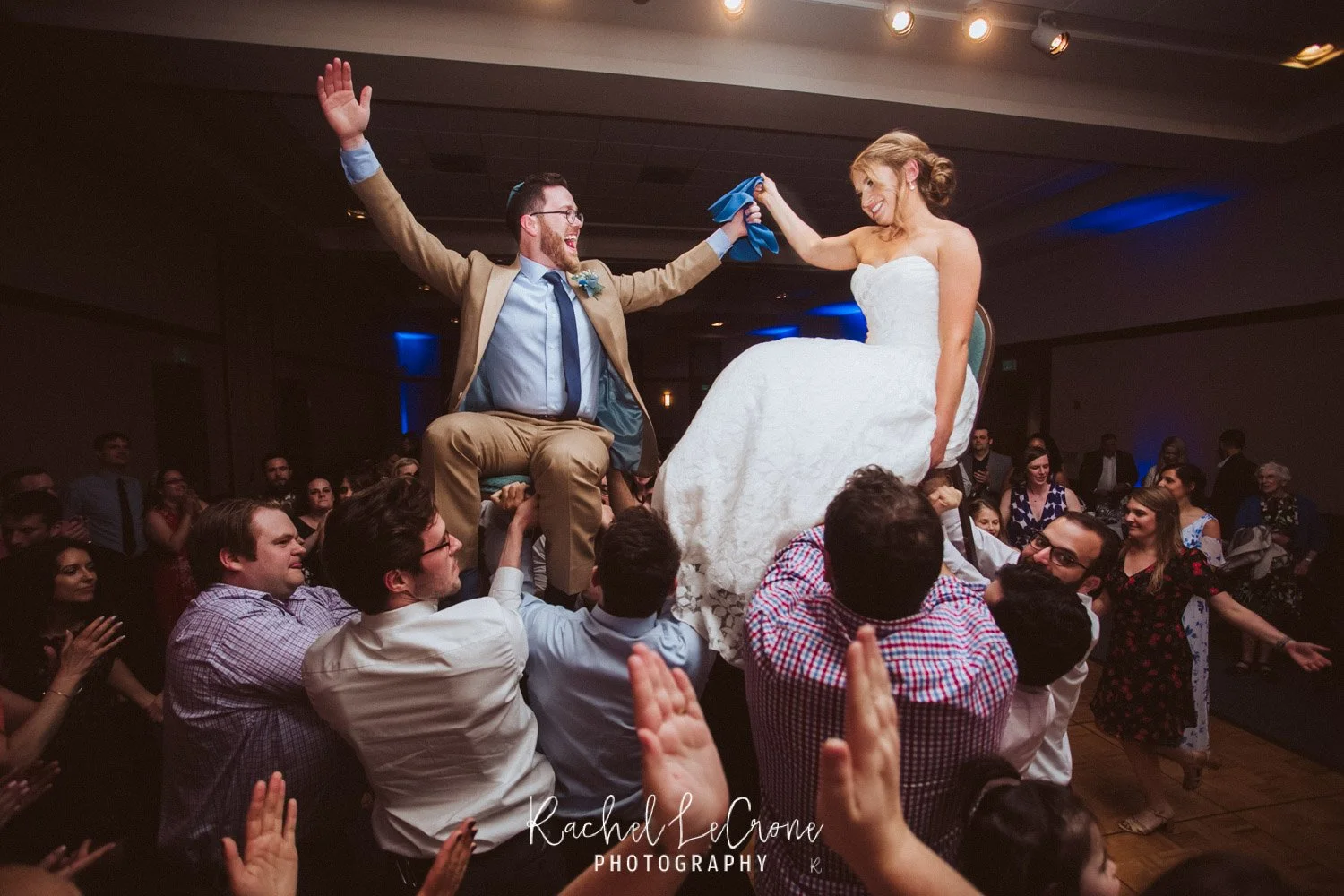 A bride and groom sitting on chairs lifted above the crowd at a wedding reception, smiling and celebrating as the groom holds the bride's hand; surrounded by guests clapping and cheering.