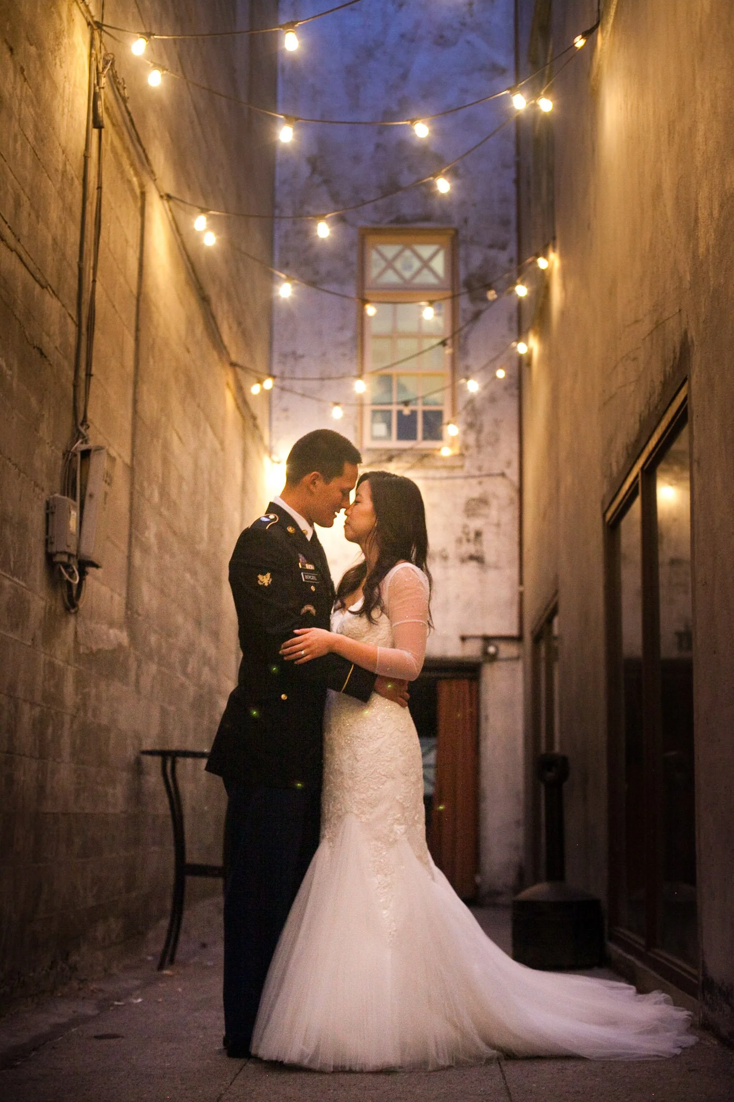 A couple in wedding attire, a man in military uniform and a woman in a white wedding gown, standing close and touching foreheads in a narrow alley decorated with string lights at night.