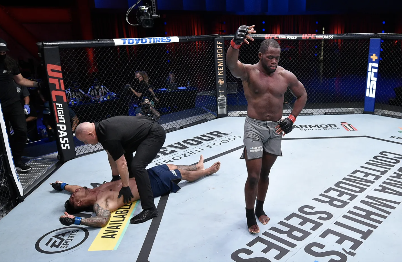 Tafon standing inside the octagon with a victorious expression, while another fighter lies on the ground with a referee checking on him after a match.