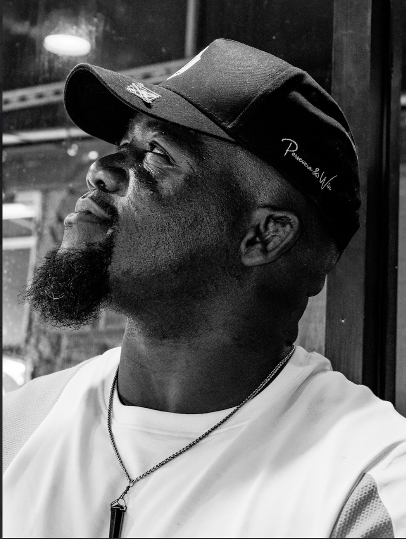 Tafon wearing a baseball cap and a white shirt, looking upward with a slight smile, in black and white
