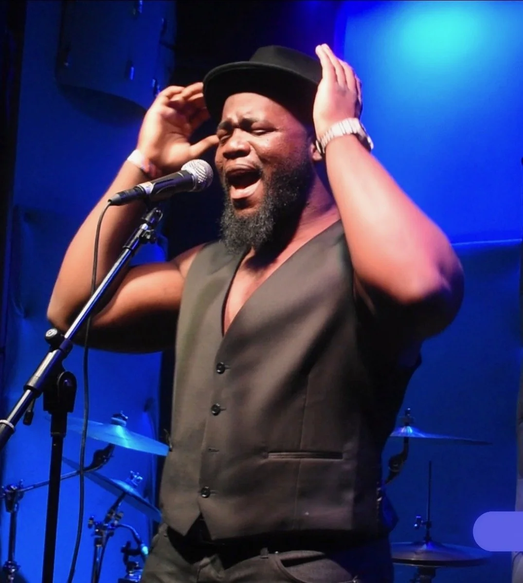 Tafon wearing a black hat, sleeveless shirt, and a watch, passionately singing into a microphone on stage with blue lighting.