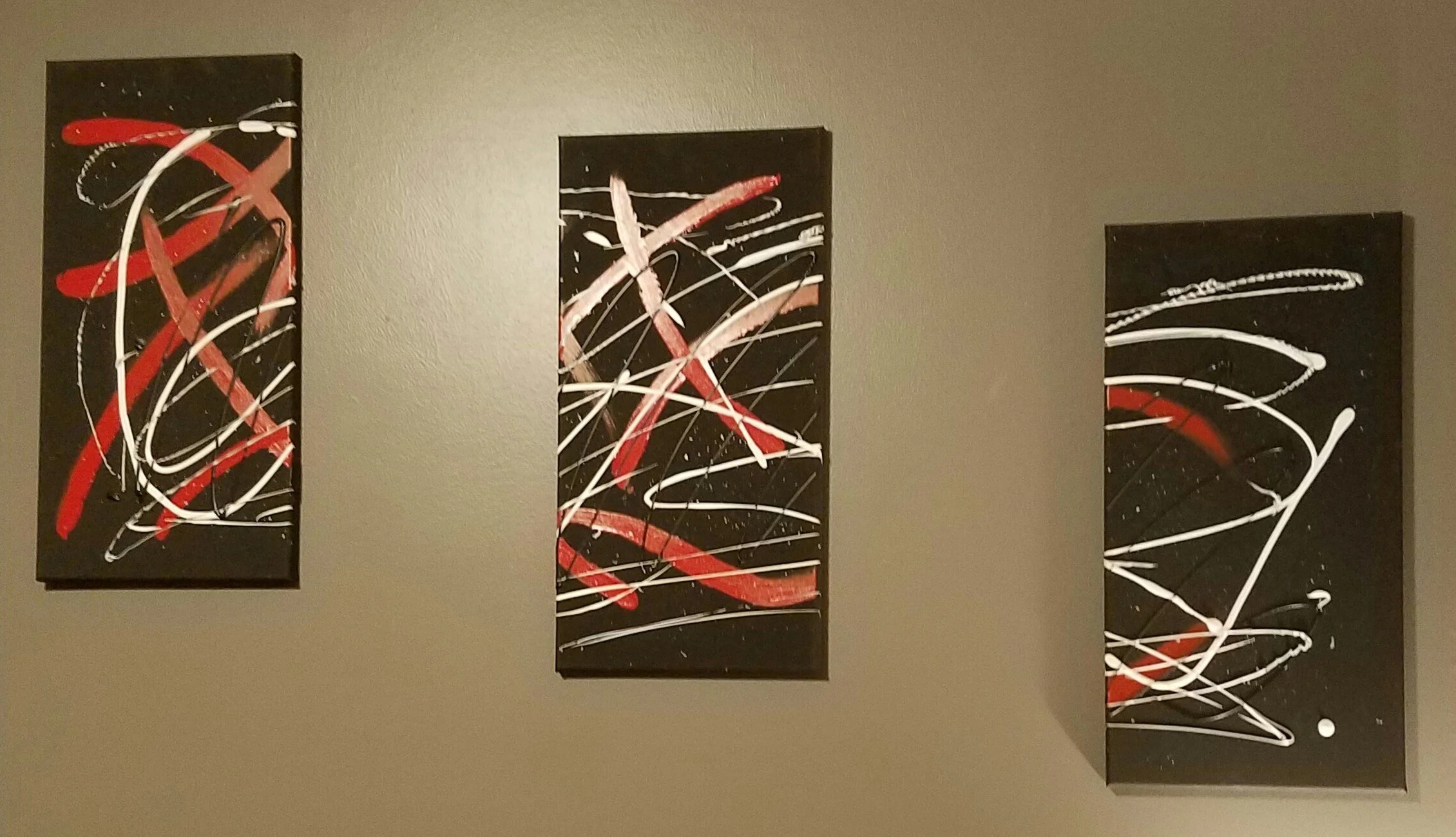 Three abstract paintings with black backgrounds and red, white, and pink streaks and lines, hung on a beige wall.