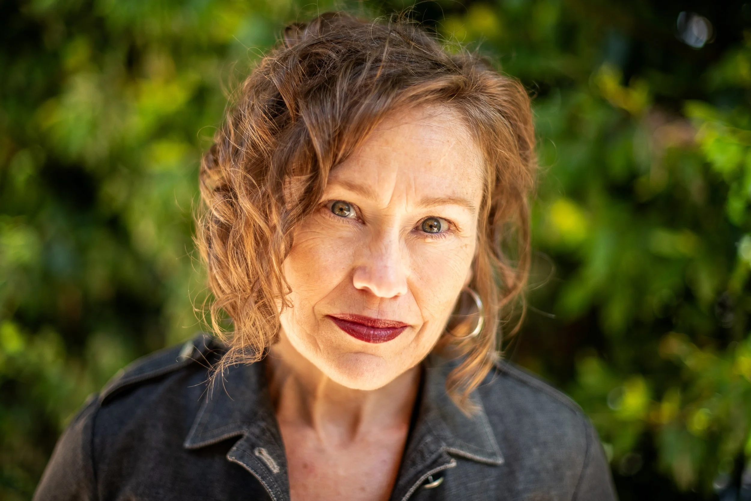 Close-up of a woman with wavy auburn hair, wearing dark lipstick, wearing a dark denim jacket, outdoors with blurred greenery background.