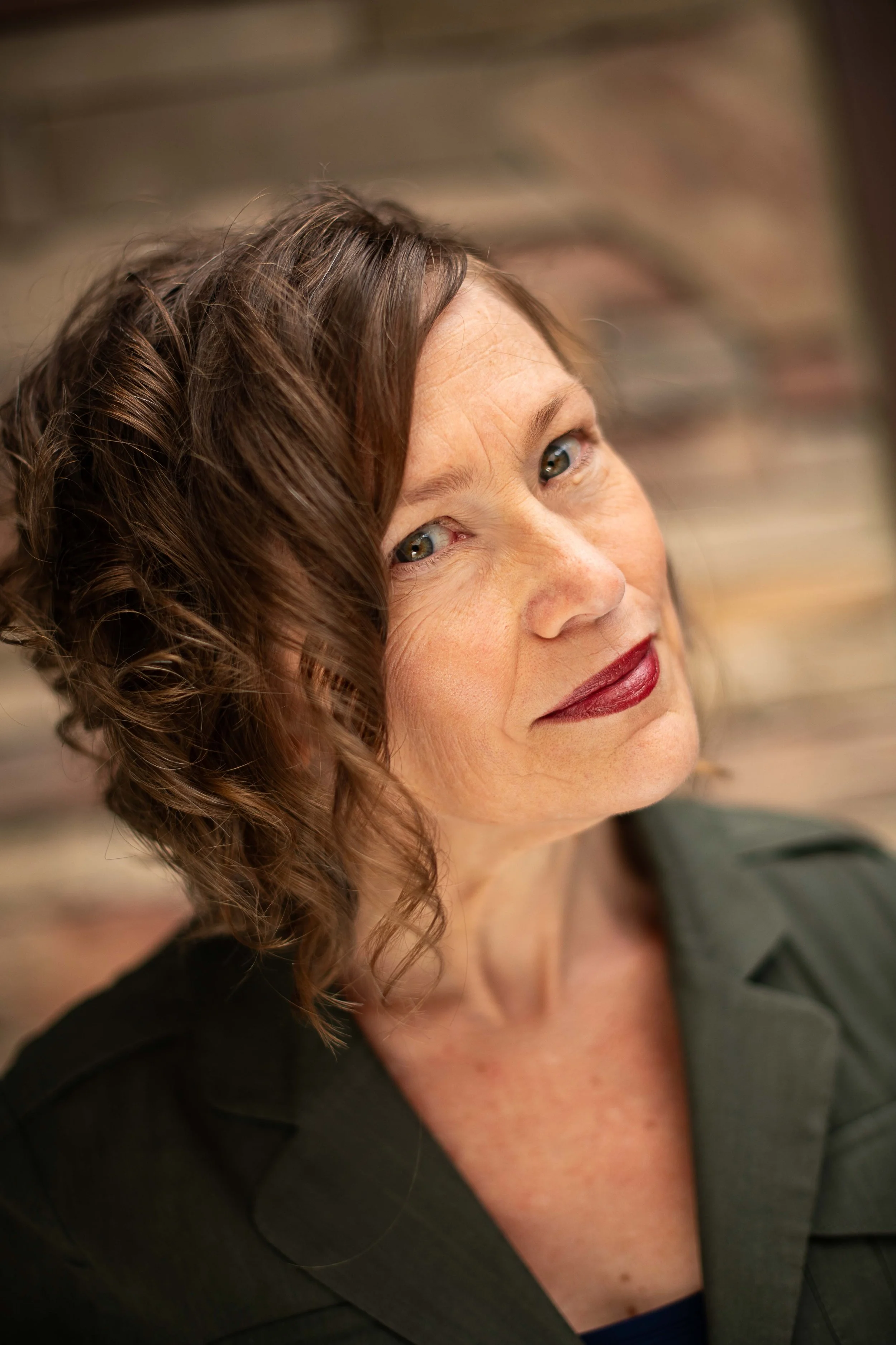 A woman with shoulder-length curly brown hair and wearing a dark blazer, looking slightly to the side with a subtle smile, in front of a blurred brick wall background.