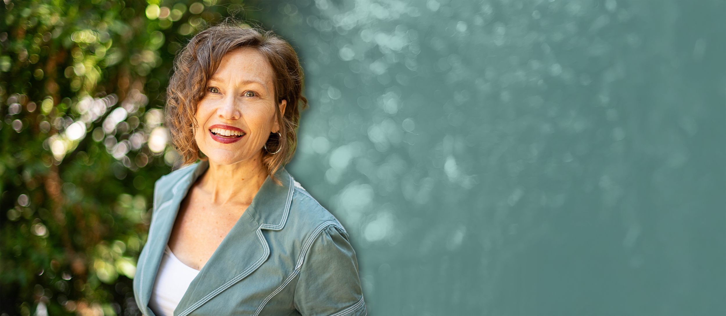 A smiling woman outdoors with green foliage background, wearing a light blue jacket and a white top.