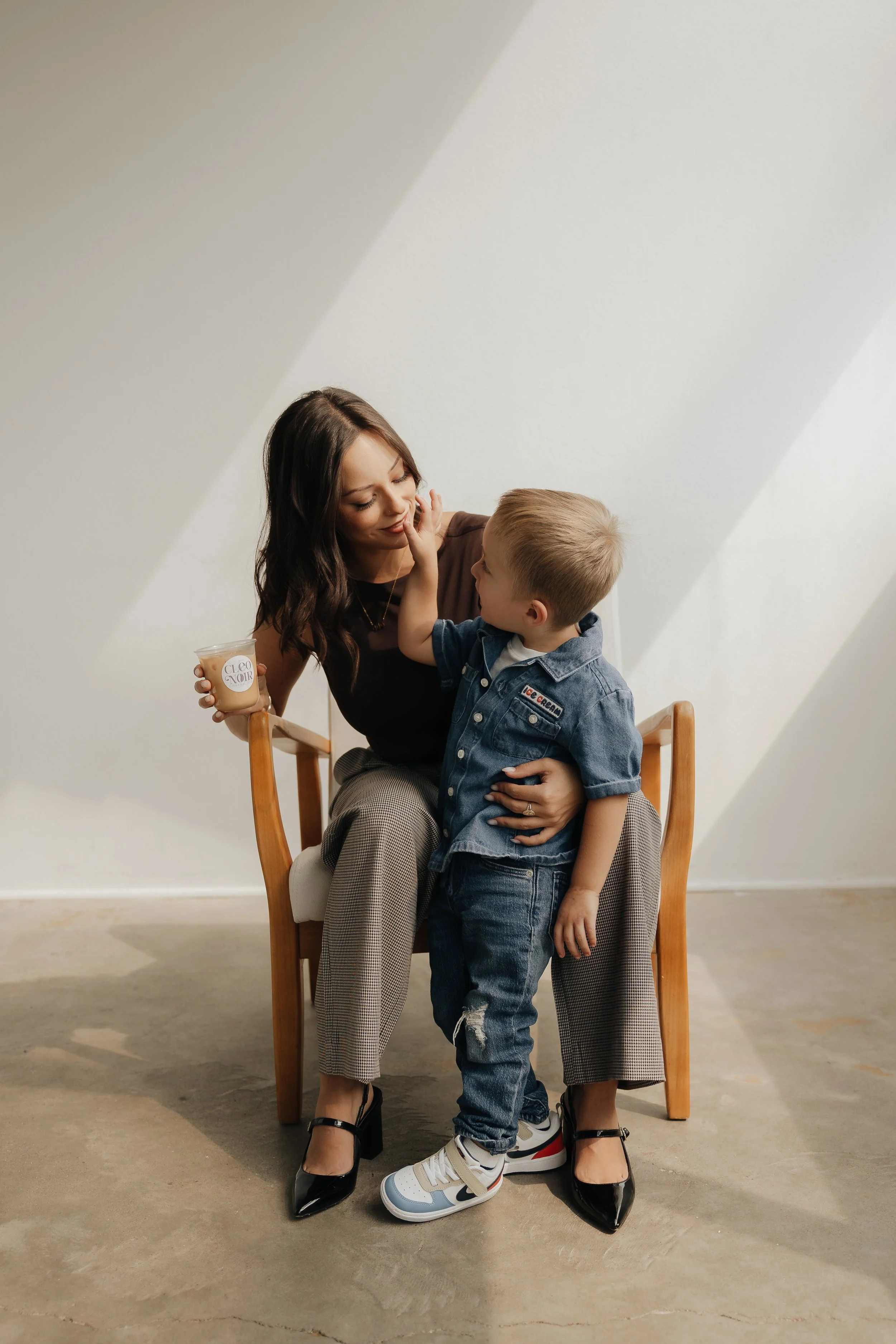 A woman and a young boy sitting on a wooden chair against a plain white wall. The woman is holding a cup of coffee labeled 'Cleo Noir' and is smiling as the boy touches her face. The boy is wearing a denim jacket, jeans, and sneakers, and the woman is dressed in black heels and checkered pants.