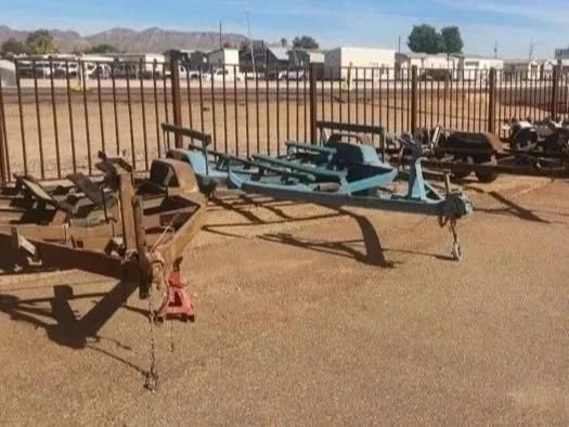 Multiple damaged boats on trailers in a fenced outdoor lot with a clear sky and buildings in the background.