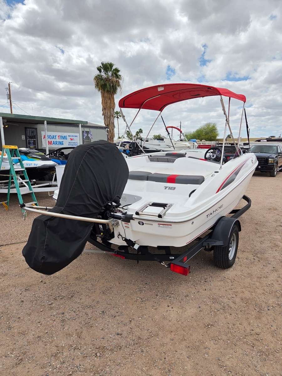 A white Tahoe boat on a trailer with a red canopy, parked on a gravel lot with palm trees and other boats and vehicles in the background.