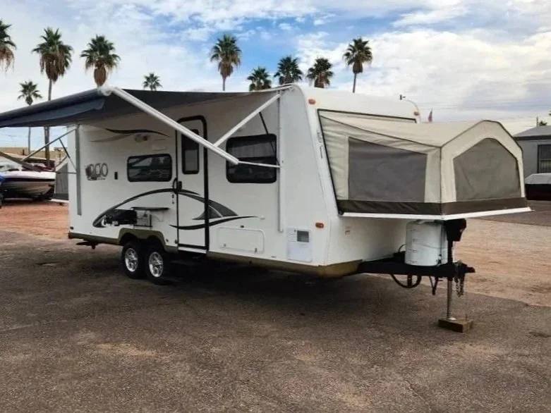 A white travel trailer with a pop-up tent attached at the front, parked on a gravel lot with palm trees in the background.