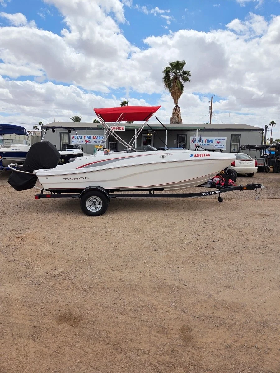 A white Tahoe motorboat on a single-axle trailer parked on a gravel lot, with a black cover over the engine, a red canopy, and a palm tree in the background under a partly cloudy sky.