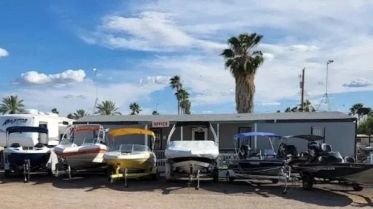Row of small boats on trailers parked in a lot with palm trees and a blue sky in the background.