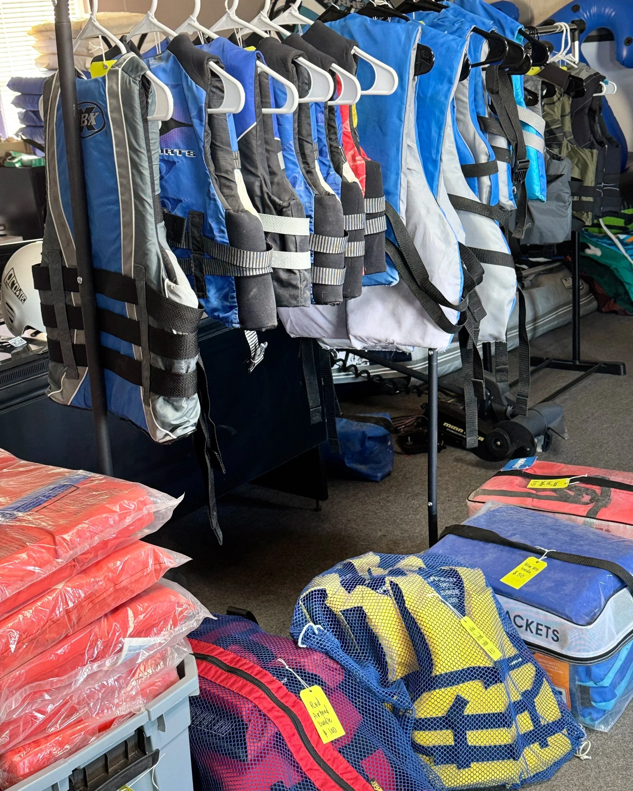 A display of blue, black, and white life jackets hanging on a rack, with orange life jackets and yellow and blue bags on the table below.