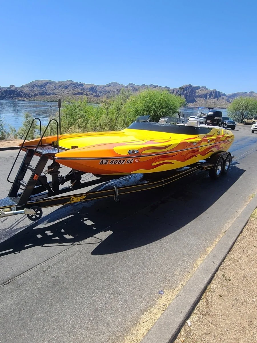A race boat with a flame design on a trailer, parked on a road near a lake with mountains in the background.