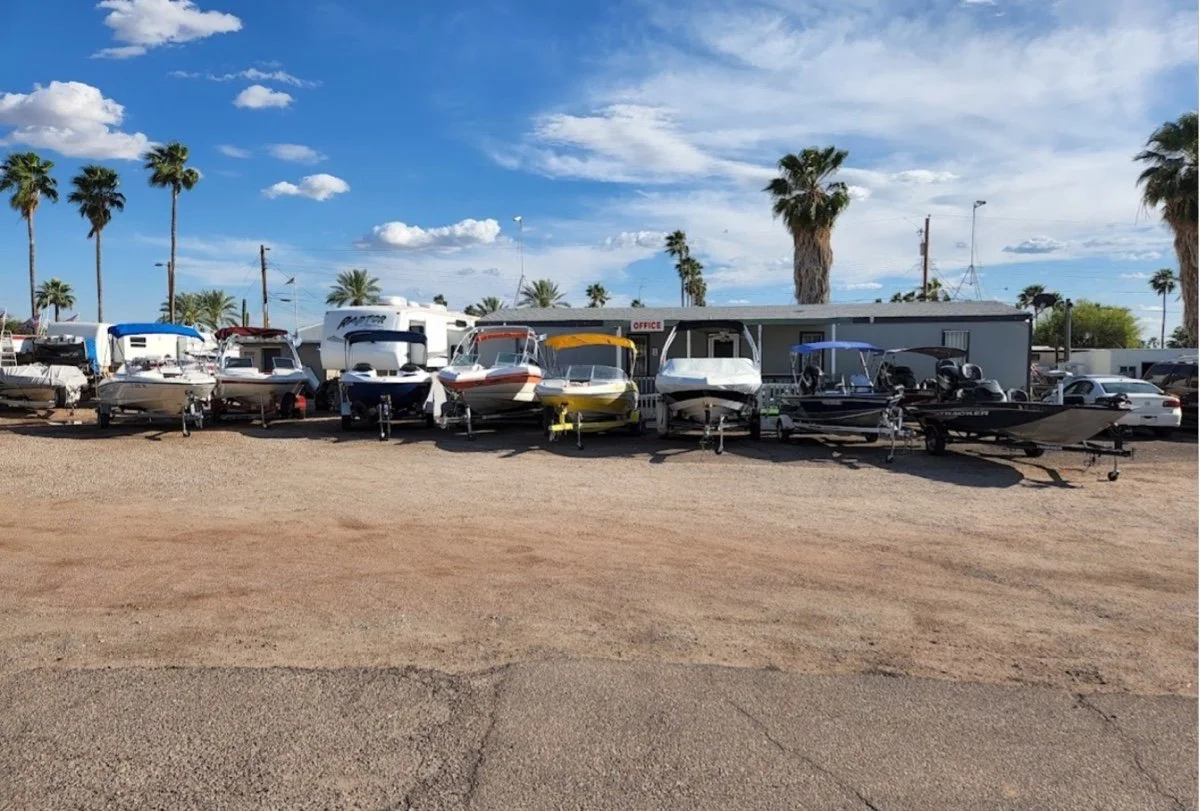 A boat dealership lot with multiple boats on trailers parked in front of a small office building against a backdrop of tall palm trees and a partly cloudy blue sky.