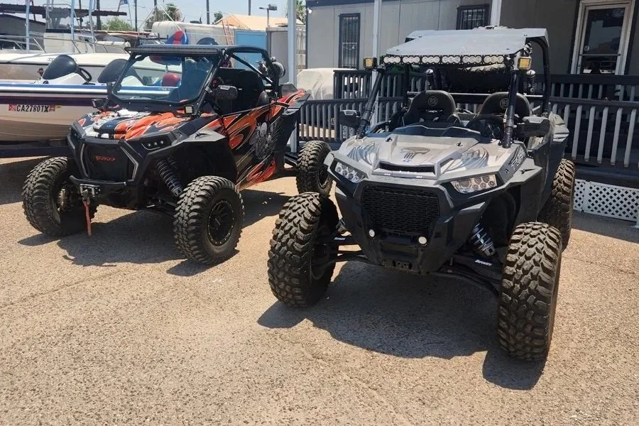 Two off-road utility terrain vehicles (UTVs) parked on a gravel surface, with boats and a building in the background.