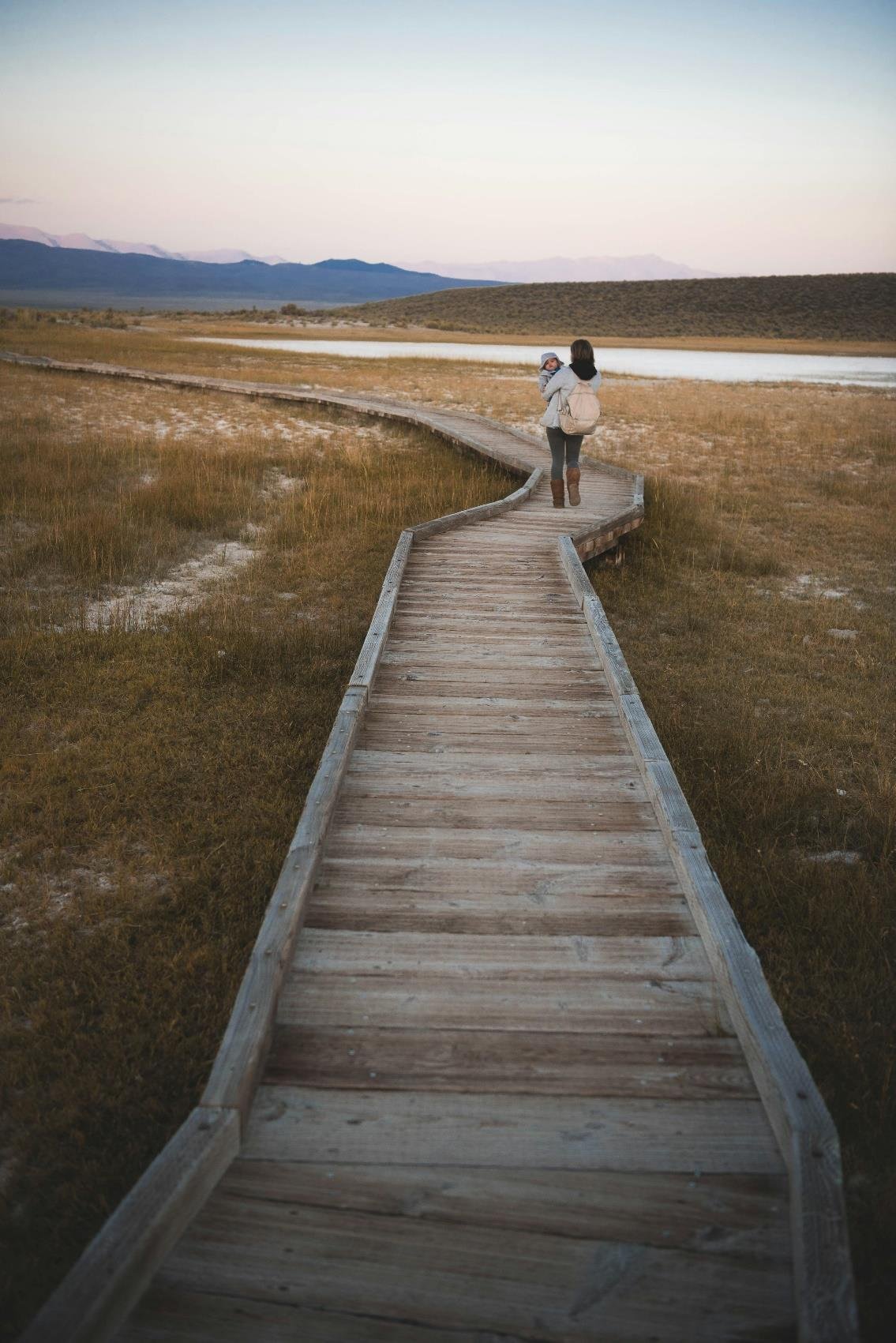 A woman walking on a winding wooden boardwalk through a dry, grassy landscape with a woman carrying a child, near a body of water, mountains in the background, during possibly sunset or sunrise.