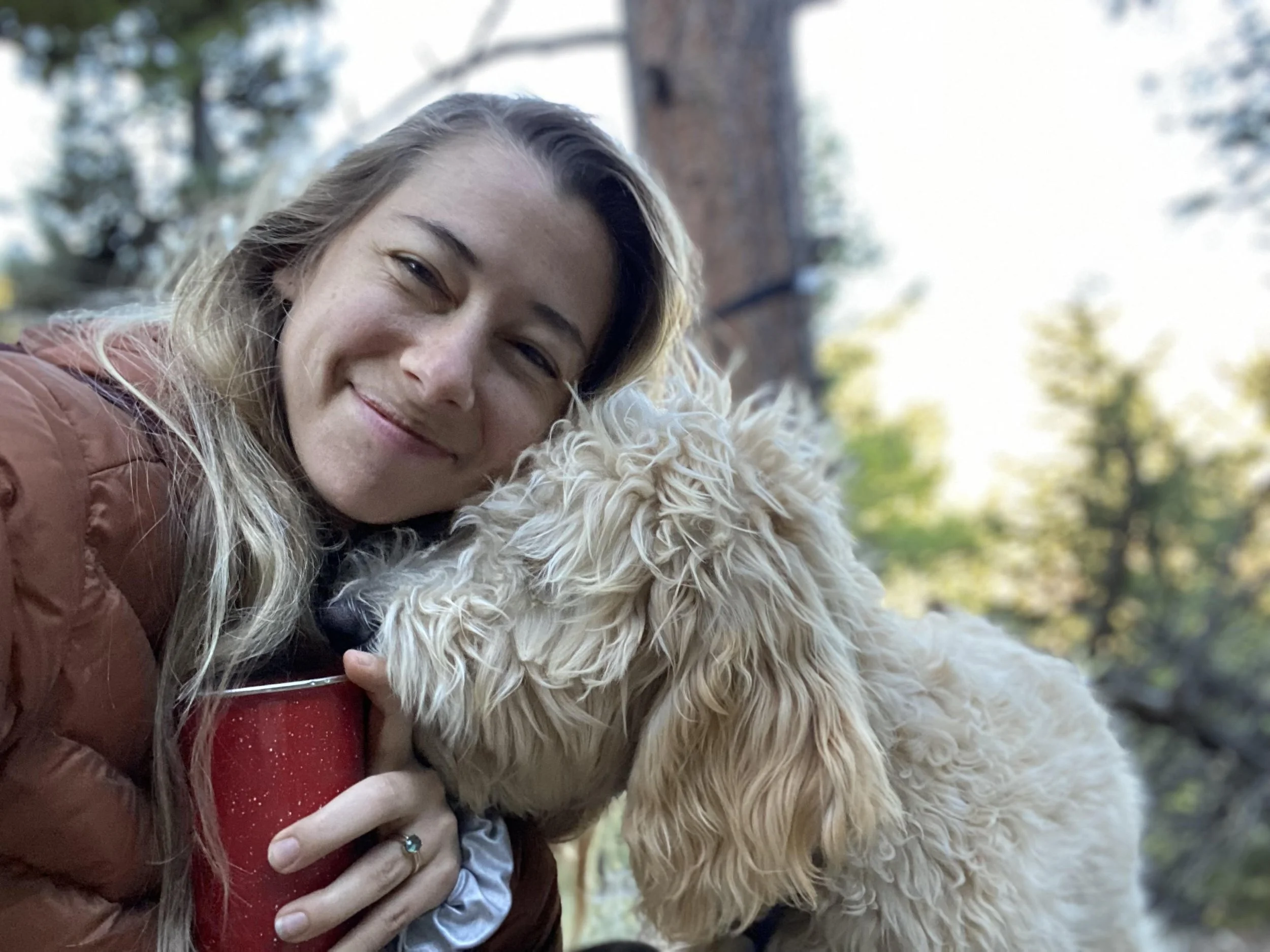 A woman smiling and holding a red cup next to a curly-haired dog outdoors in a wooded area with trees and sunlight in the background.