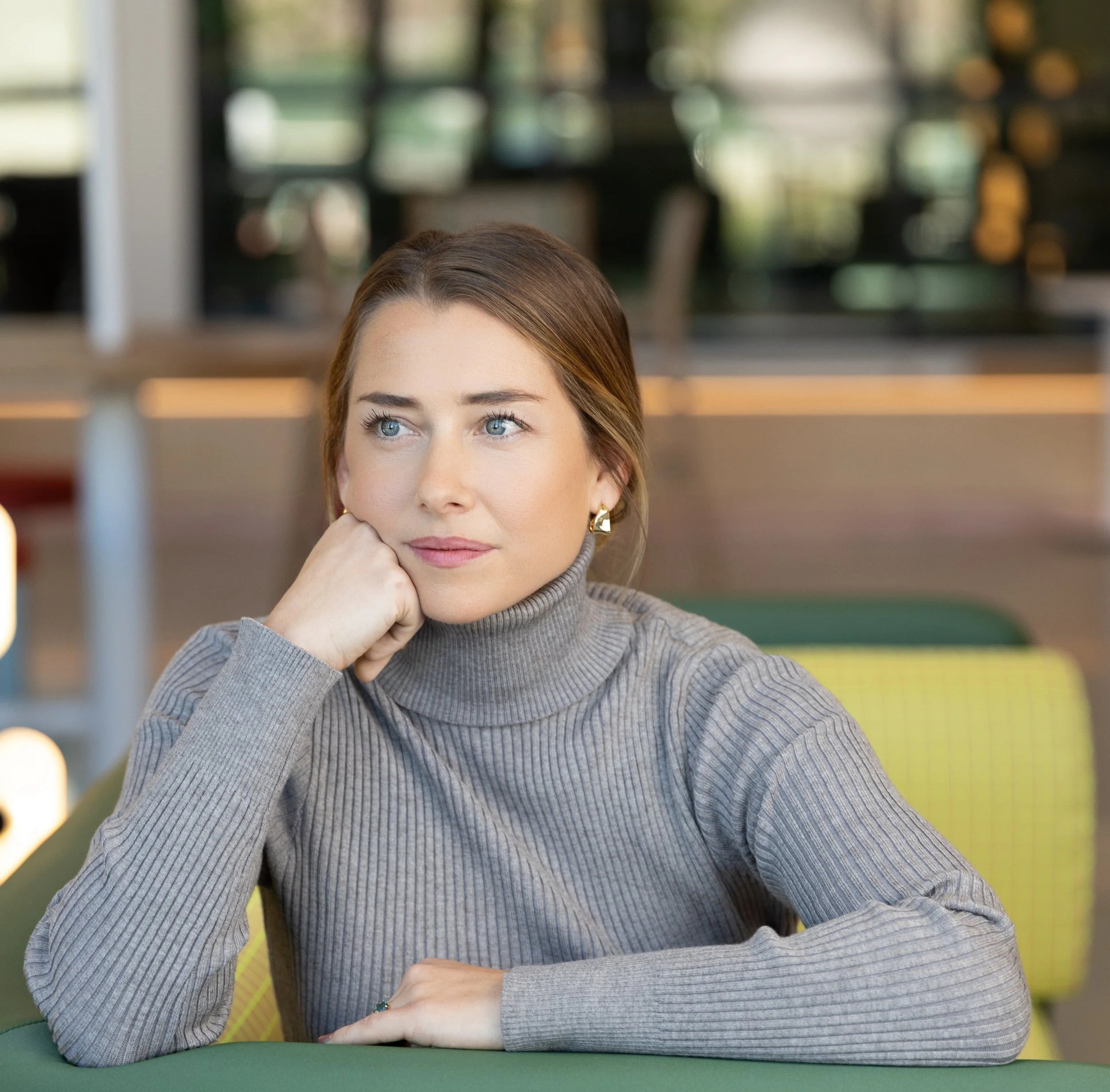 A woman in a gray turtleneck sweater sitting at a table with her head resting on her hand, looking pensively out of frame in a well-lit indoor space.
