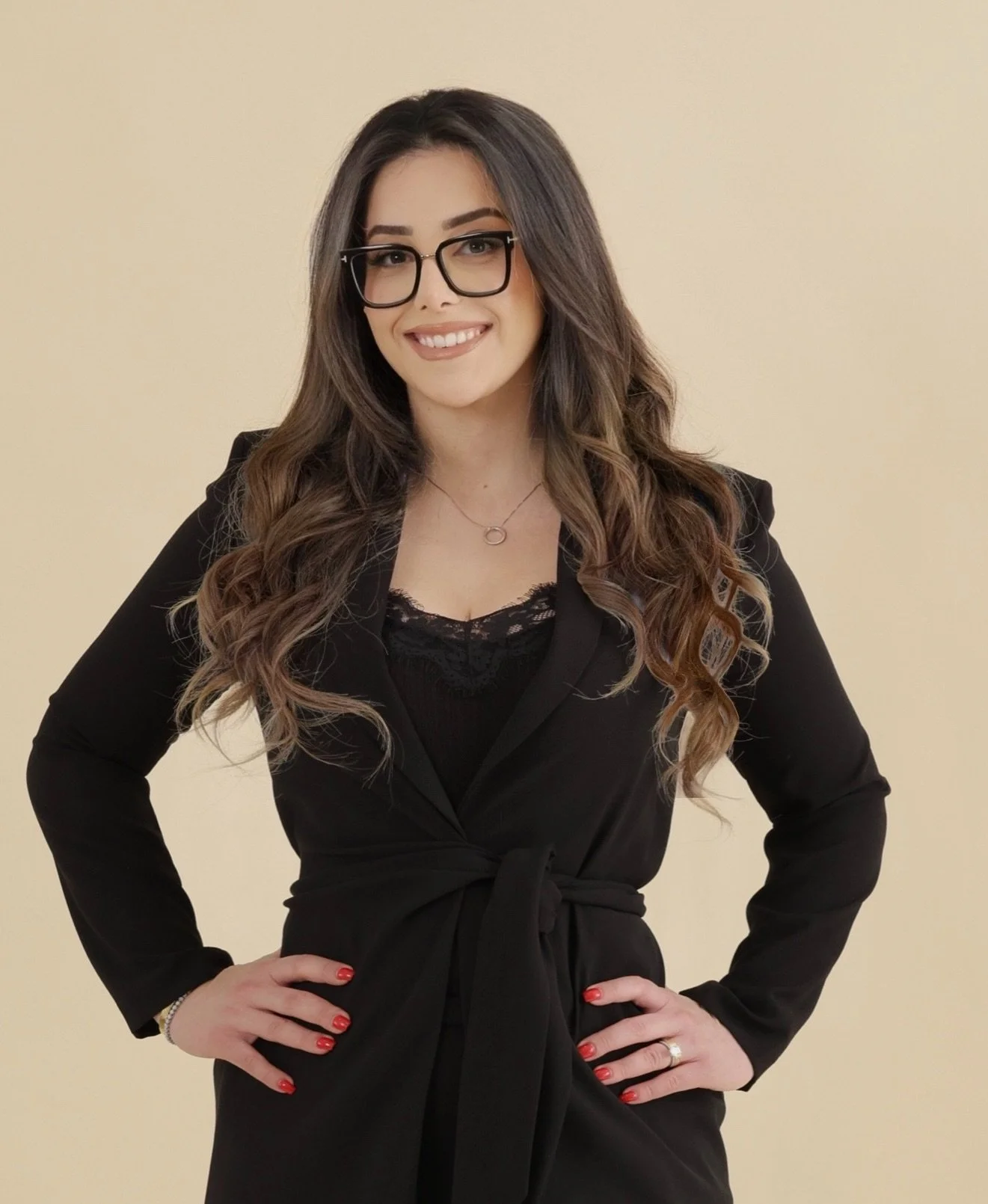 A woman with long, wavy brown hair, wearing a black blazer and glasses, standing with hands on hips, smiling, against a plain beige background.