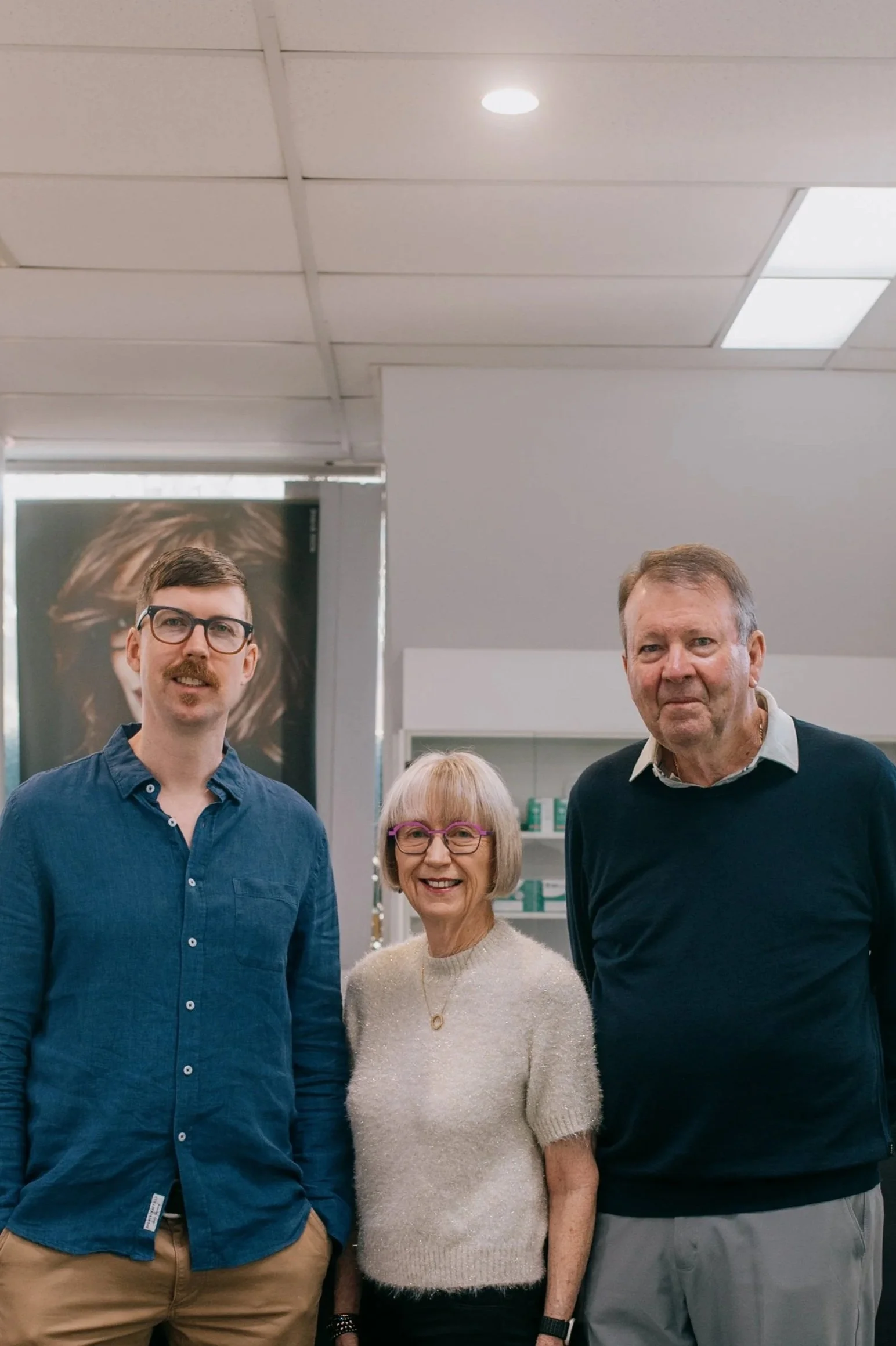 Three people standing indoors, smiling at the camera, with a cabinet and a poster of a person with red hair in the background.