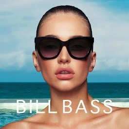 Woman with pulled-back hair wearing large sunglasses, standing outdoors near the water with a clear sky in the background, and the text 'BILL BASS' at the bottom.