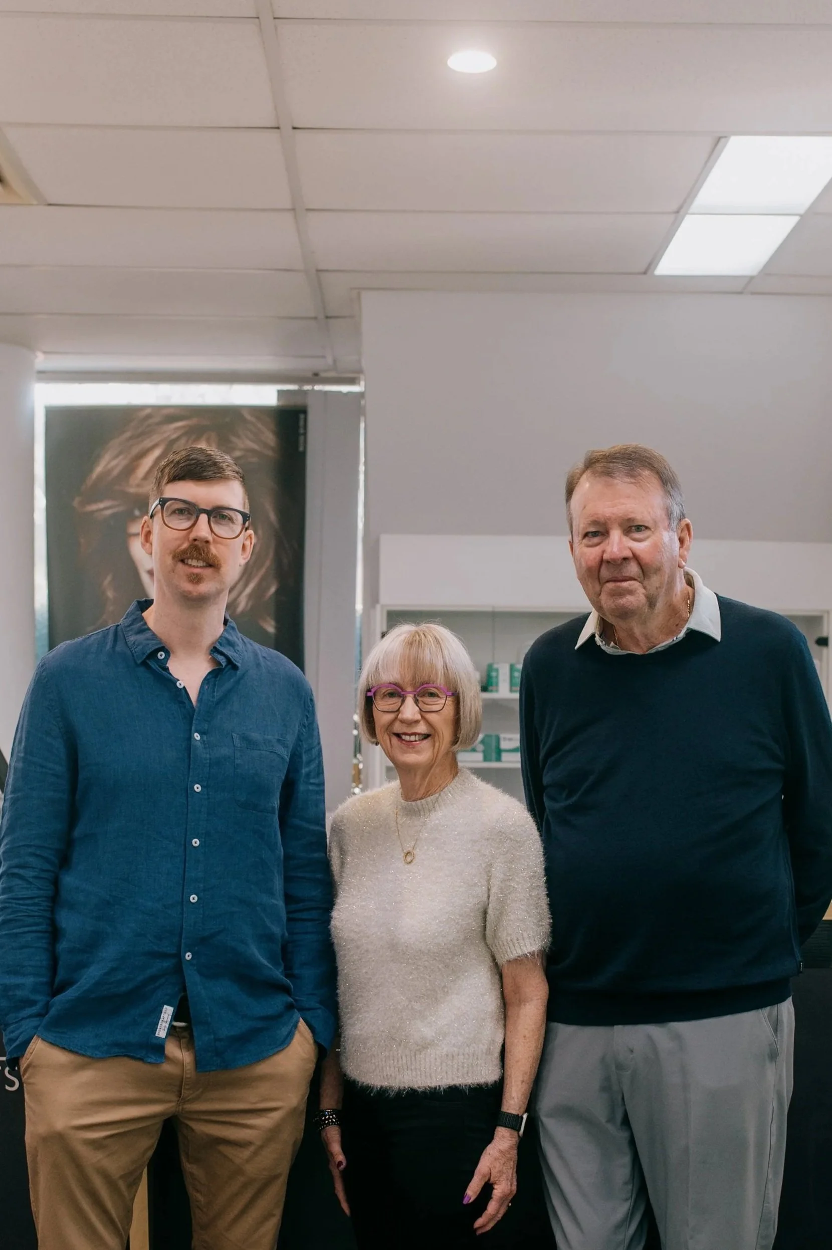 Three people standing together indoors, smiling at the camera. A young man with glasses and a blue shirt on the left, an older woman with glasses and a cream sweater in the middle, and an older man in a dark sweater on the right.