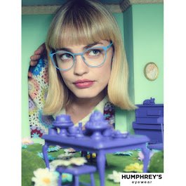 A young woman with blonde hair, wearing glasses and a colorful sweater, looking at a model of the Titanic ship in front of her in a room with mint green walls.