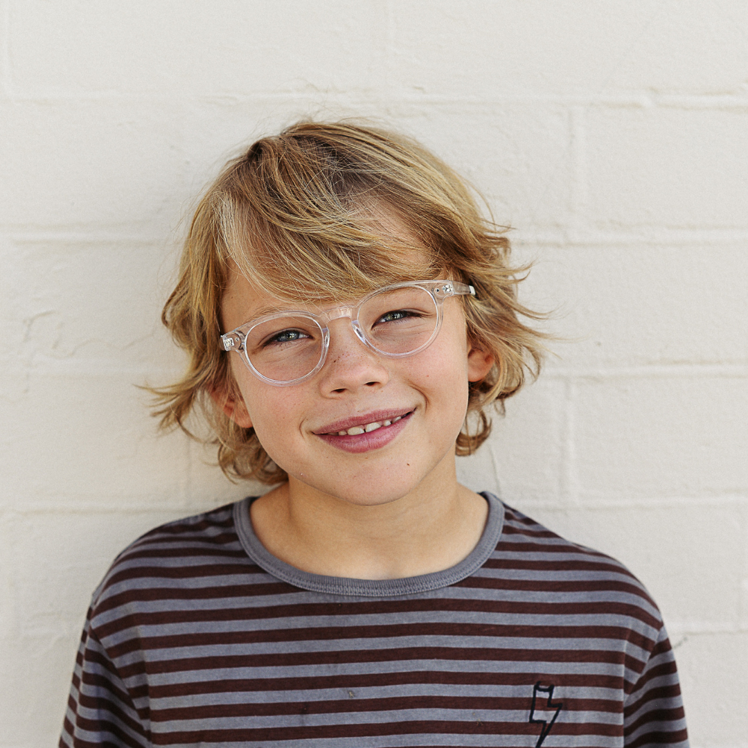 Young boy with blonde hair, wearing clear glasses and a striped gray and black shirt, smiling and standing against a white brick wall.