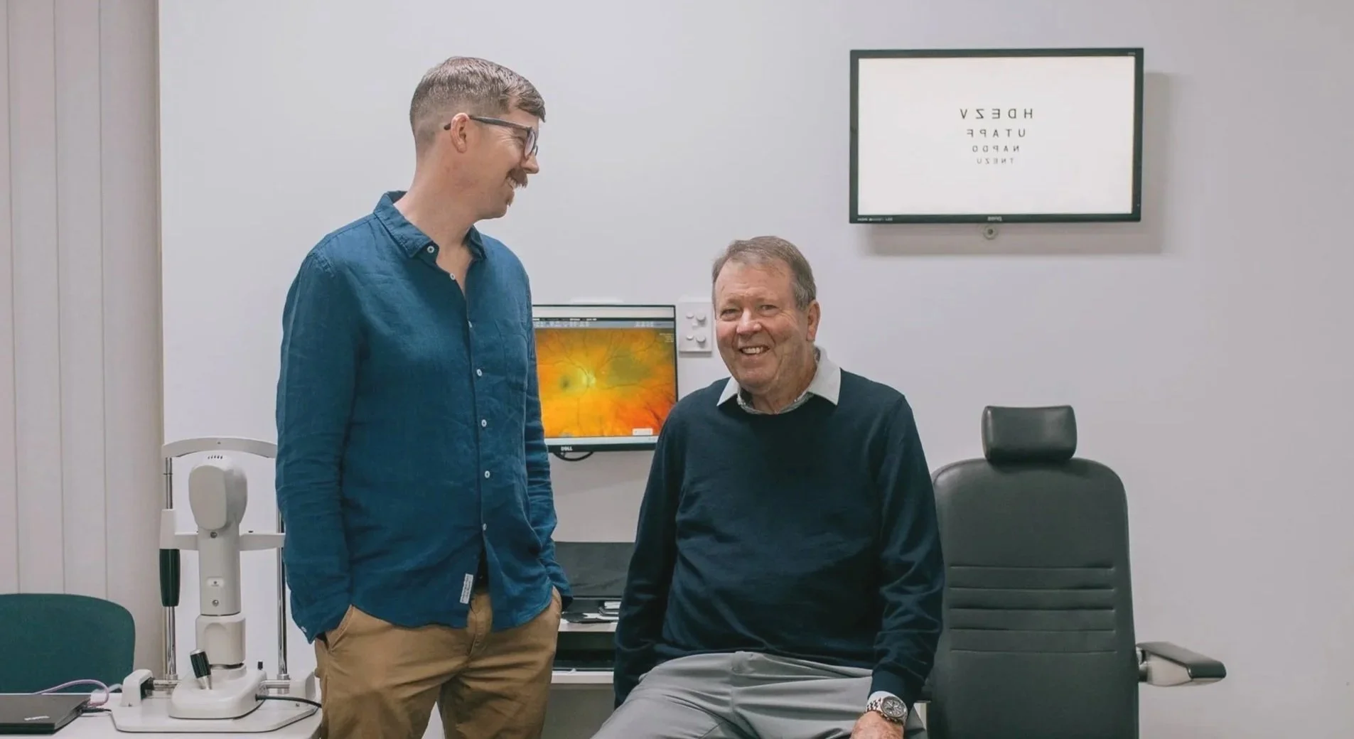 Two men in an eye doctor's office, one standing and smiling, the other sitting in a chair, with medical equipment and eyeglasses test chart on the wall.