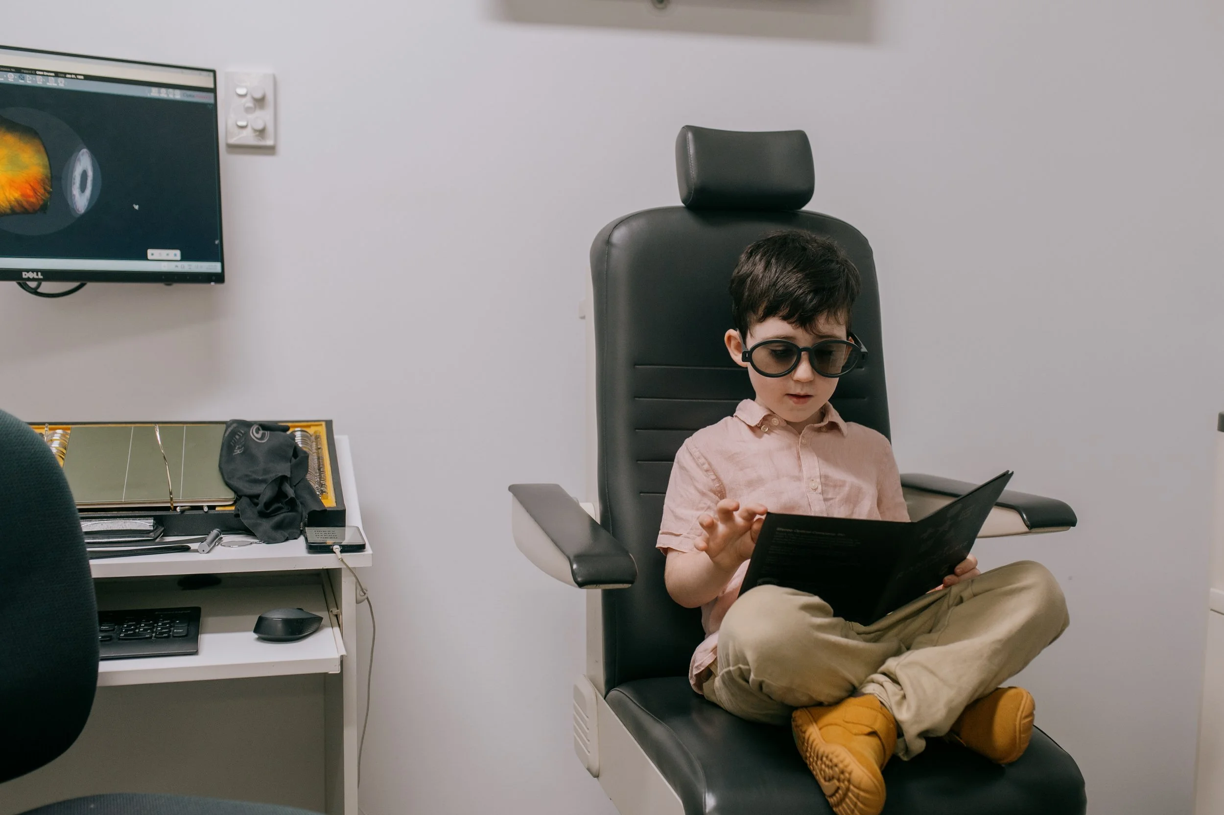A young boy wearing glasses, a pink shirt, and khaki pants is sitting in a black office chair, looking at a brochure or magazine in his hands. There is a computer monitor displaying a colorful image on the wall to his left, and a white desk with various items including a black mask, a screwdriver, and a small electronic device.