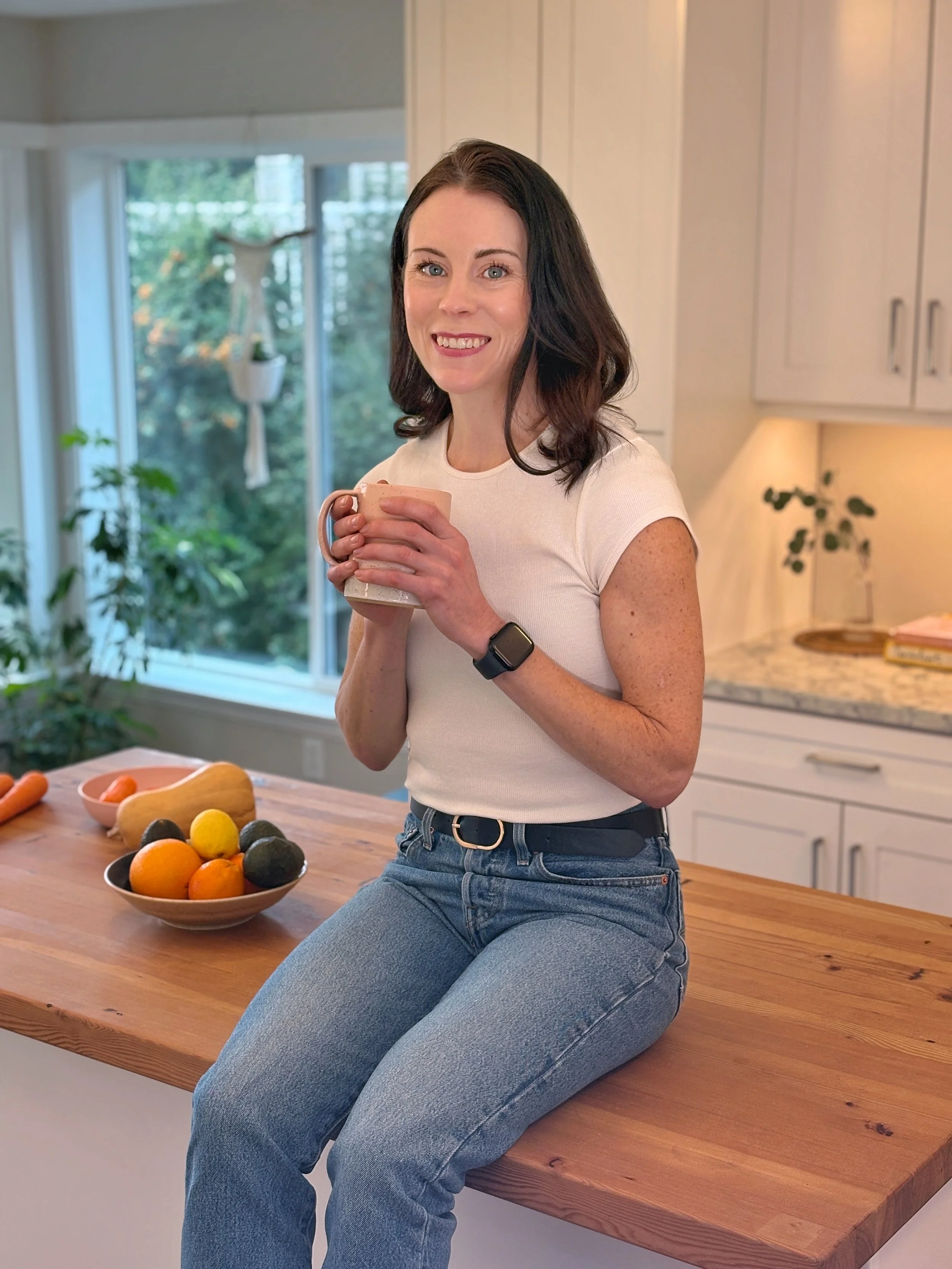 A woman with brown hair, wearing a white t-shirt and jeans, sitting on a kitchen counter, holding a mug and smiling at the camera. The kitchen has a wooden countertop, a bowl of colorful fruit, and large windows with greenery outside.