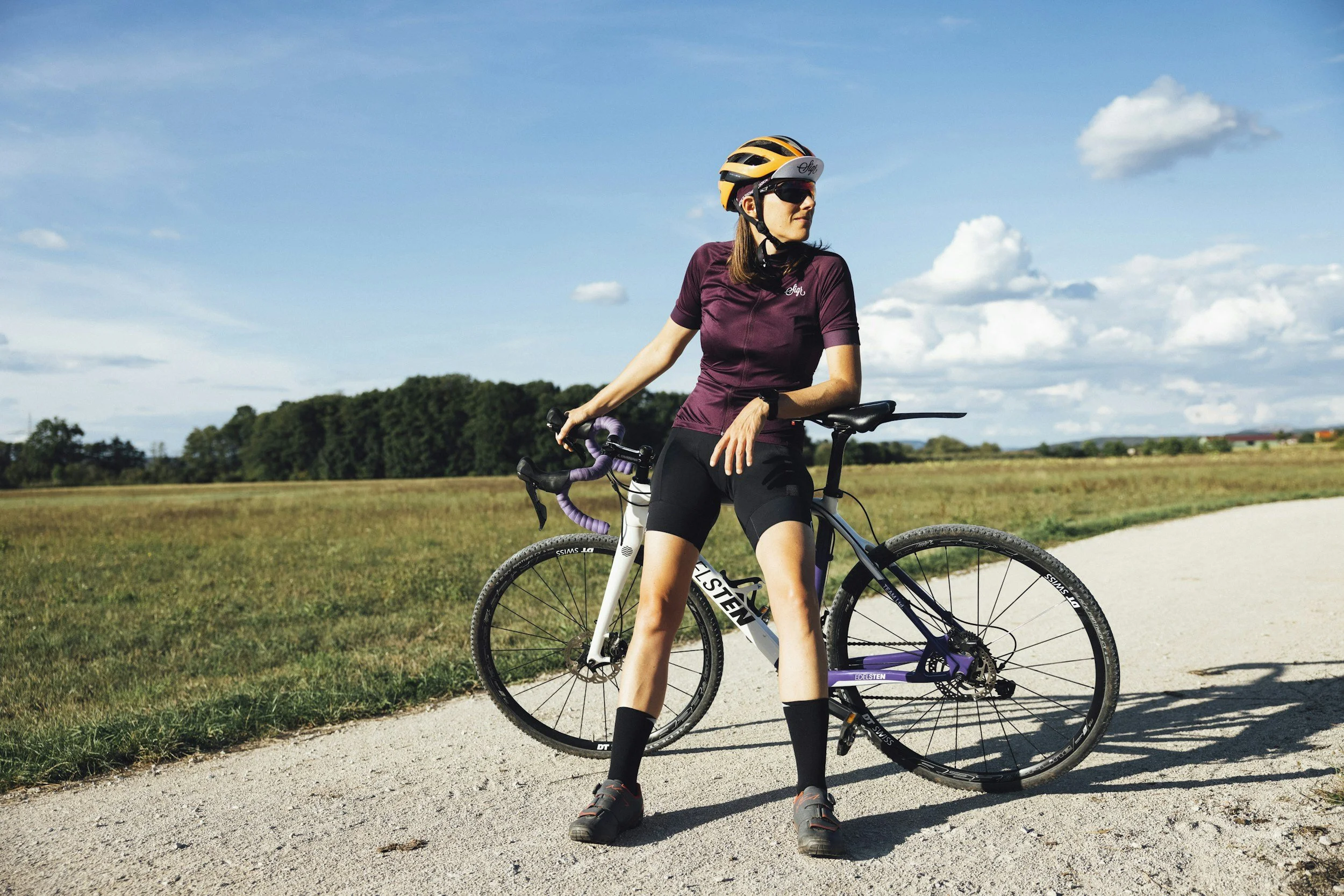 A woman wearing cycling gear and helmet standing with her bicycle on a rural path under a blue sky with clouds.