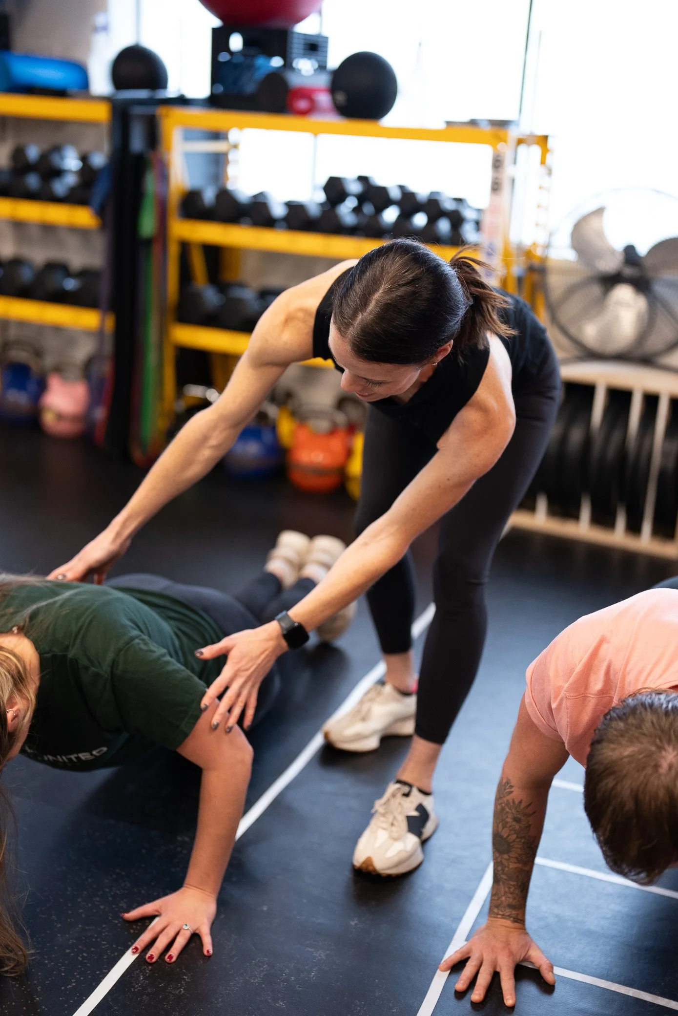 A woman in a black workout outfit assisting two people during a workout session at a gym. The gym has black flooring, yellow shelves with equipment, and large windows.