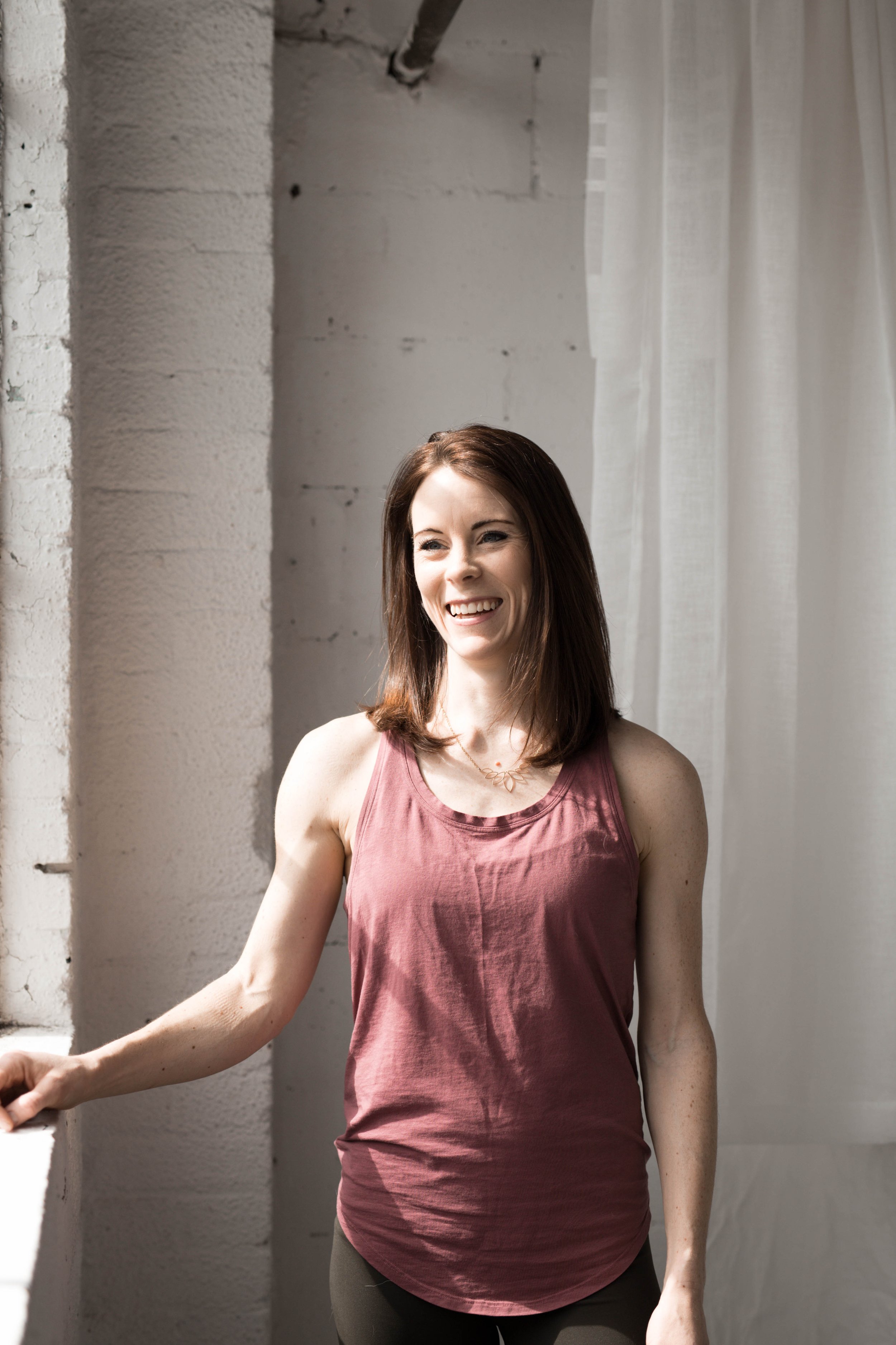 A woman with shoulder-length brown hair smiling indoors near a white brick wall and white curtains.