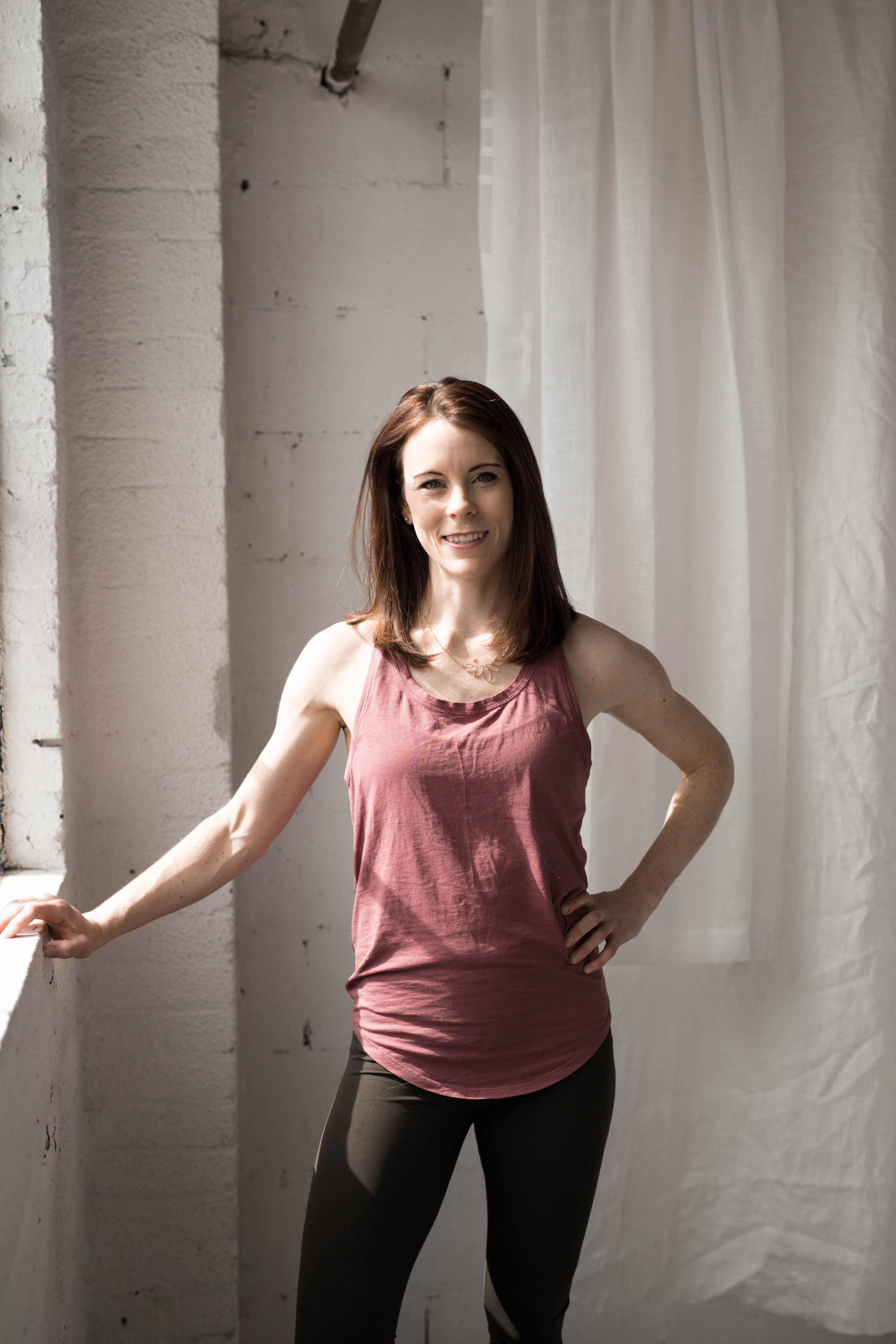 A woman with shoulder-length brown hair wearing a pink tank top and dark leggings, standing in front of a white brick wall and white curtains, smiling and posing with one hand on her hip.