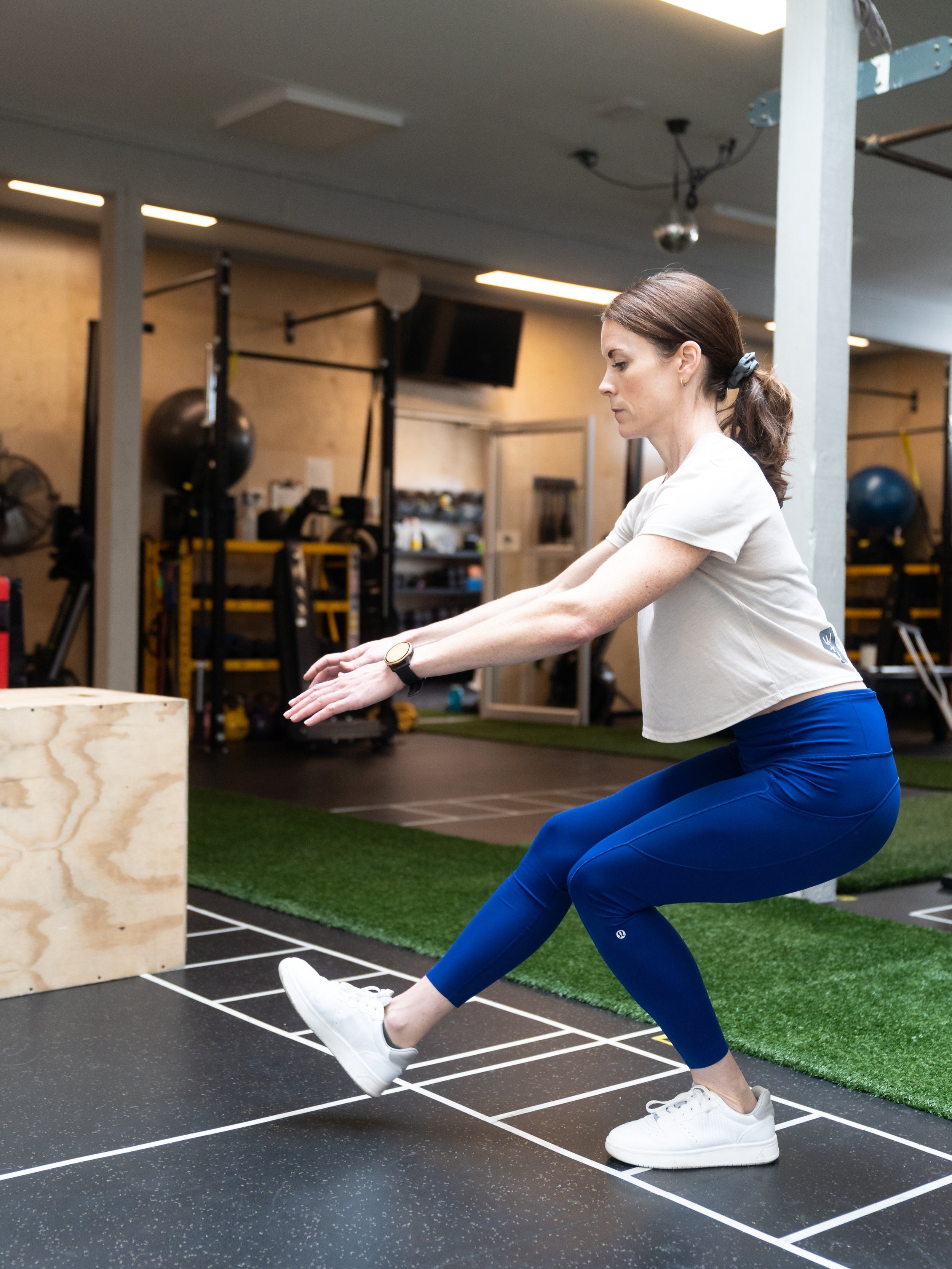 Woman doing a squat exercise in a gym, standing on black and white marked floor, wearing a white t-shirt, blue leggings, and white sneakers.