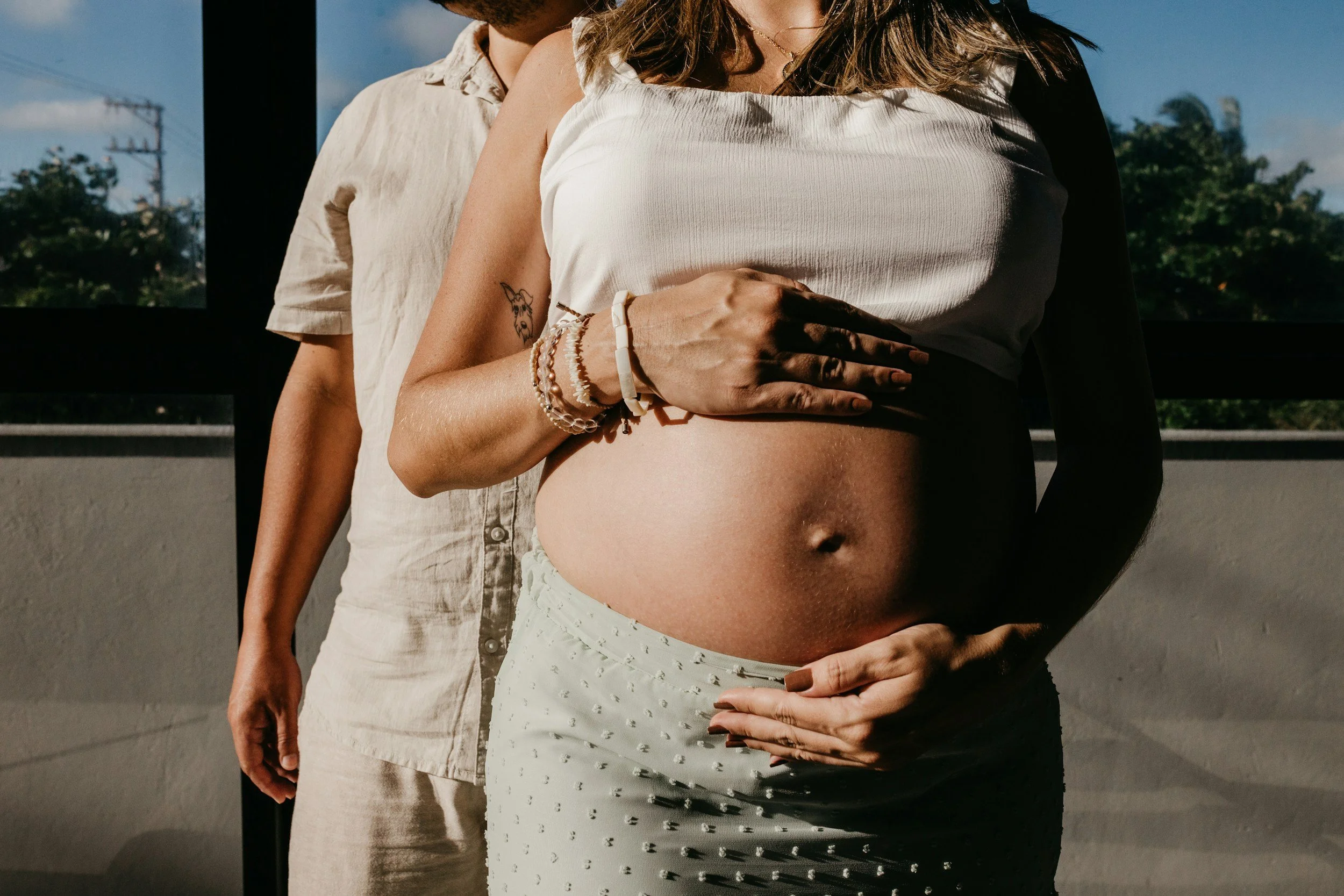 Close-up of a pregnant woman with her hand on her exposed belly, wearing bracelets, a white top, and a pale greenish blue skirt, with a man standing behind her, partly visible, in front of a window with trees reflected. The hour looks golden