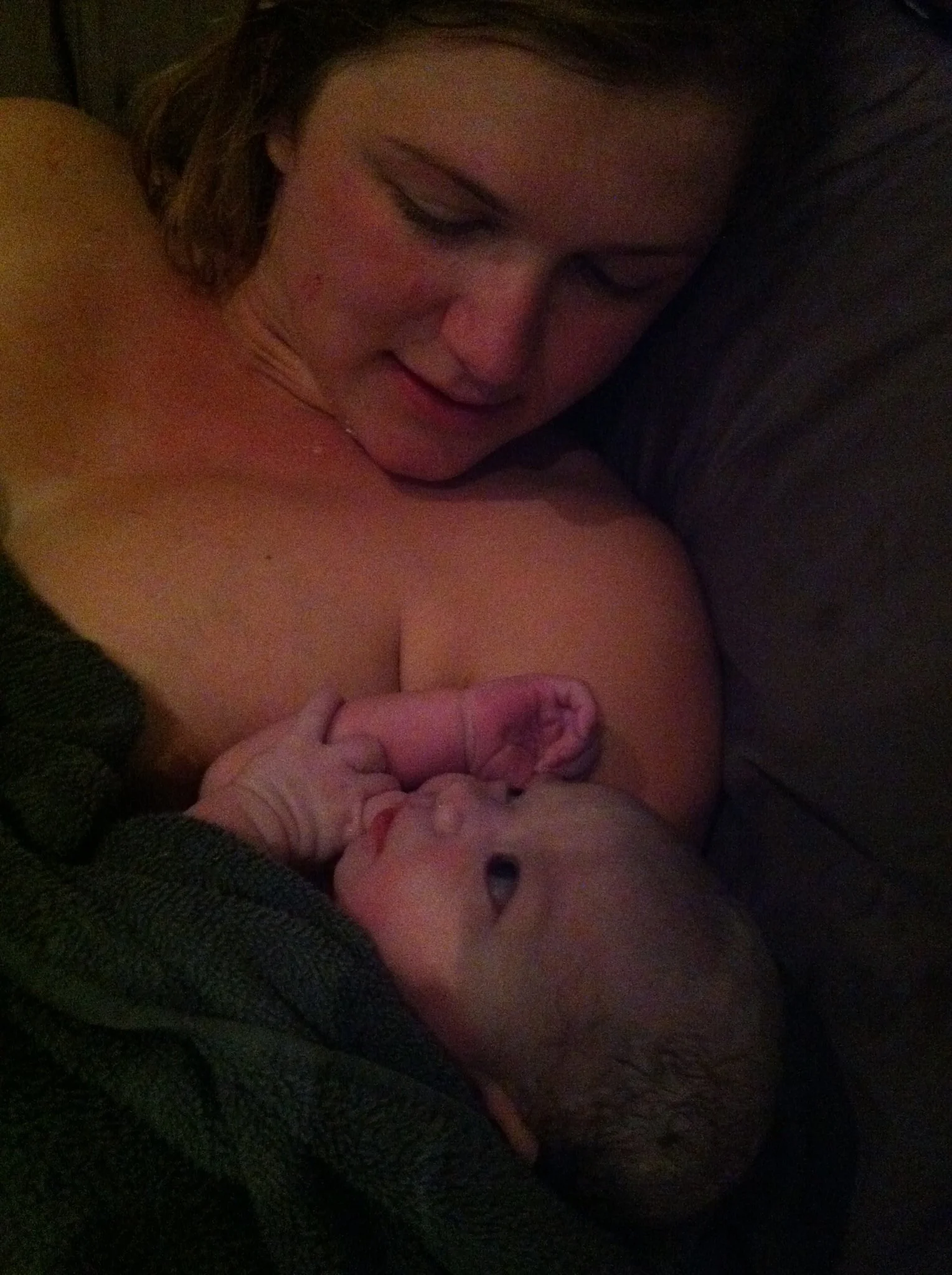 A woman with dark hair lying on a dark surface, possibly a bed or couch, holding and gently kissing a baby with light skin and dark hair.