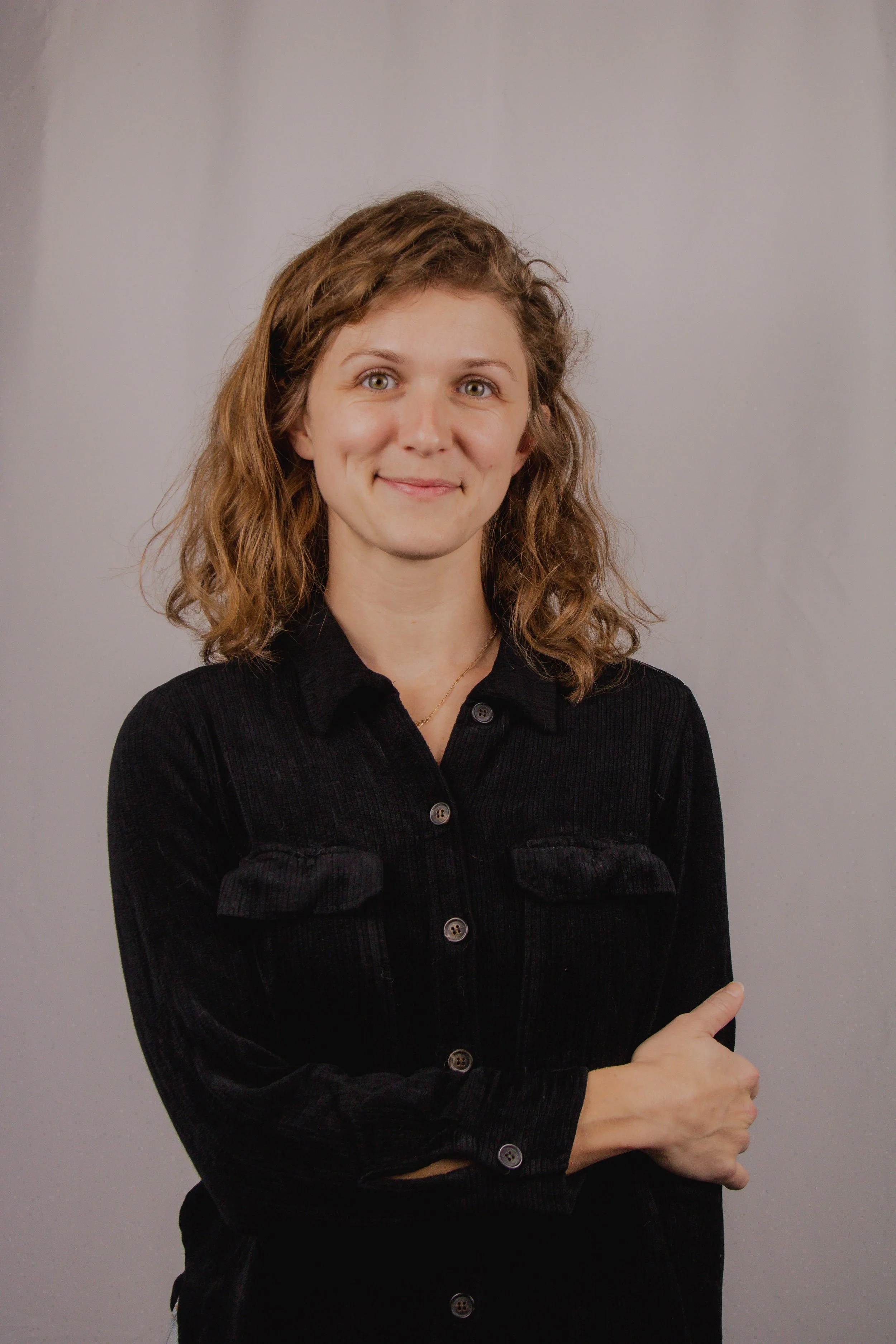 A woman with wavy brown hair wearing a black corduroy jacket, standing against a plain gray background, with her left arm crossed and pointing to the side with her right hand.