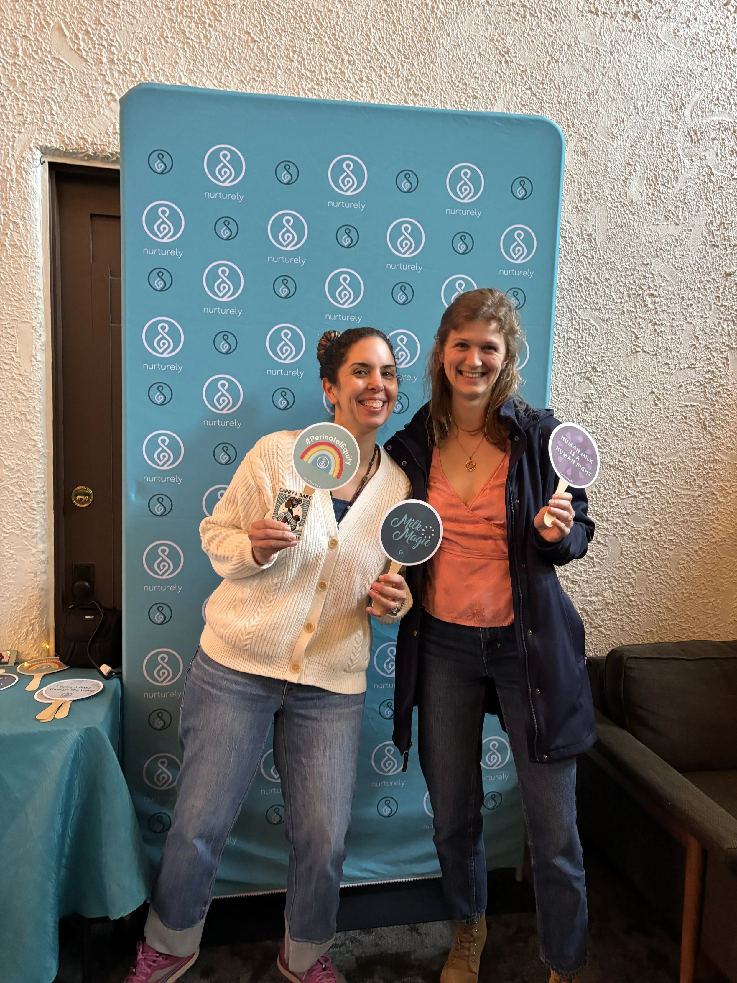 Two women smiling and holding signs at a Nurturely event, standing in front of a blue backdrop with the Nurturely logo. One woman wears a cream sweater and jeans, the other a dark jacket and jeans.