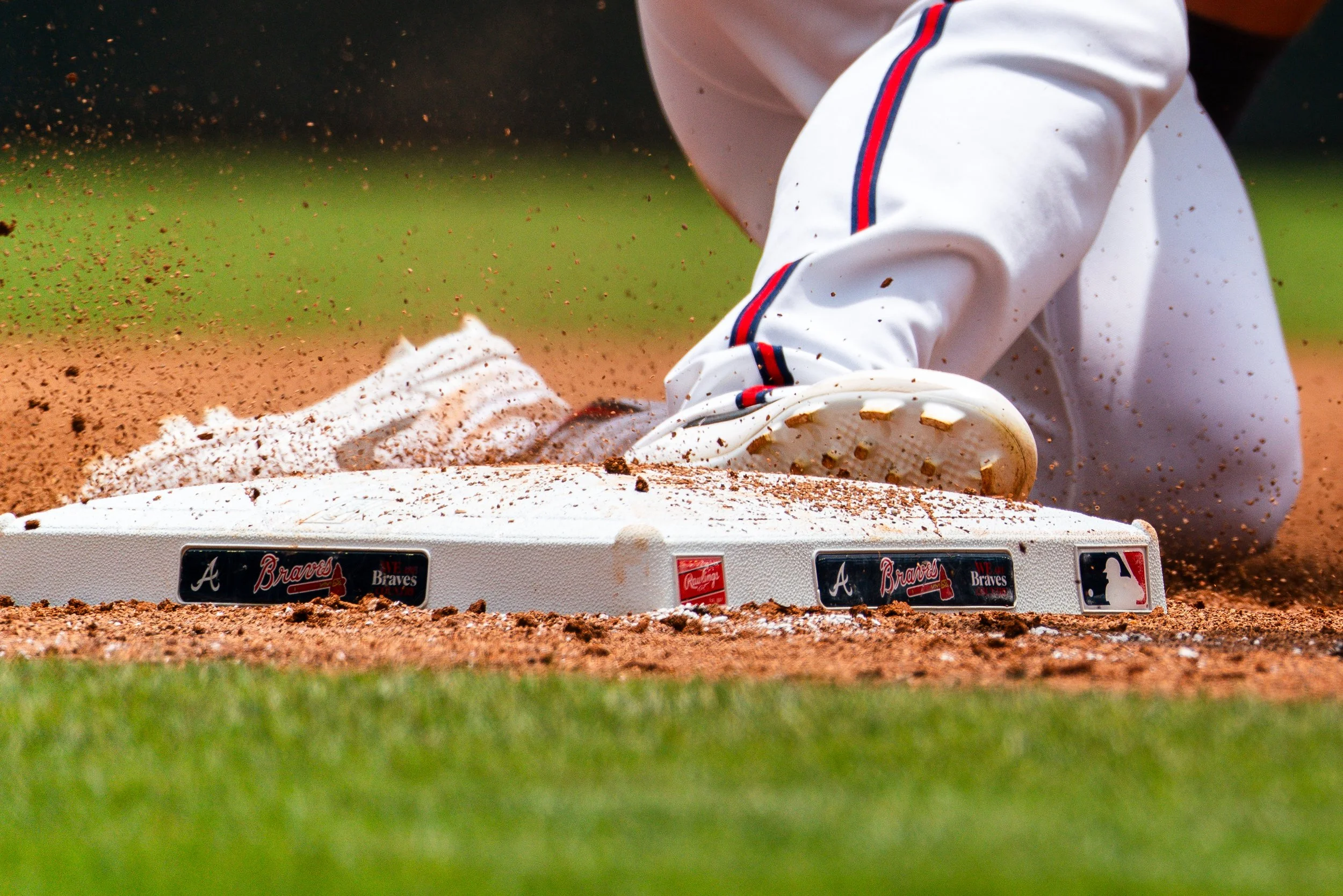 Baseball player sliding into a base with dirt flying, wearing white pants with blue and red stripes and cleats.