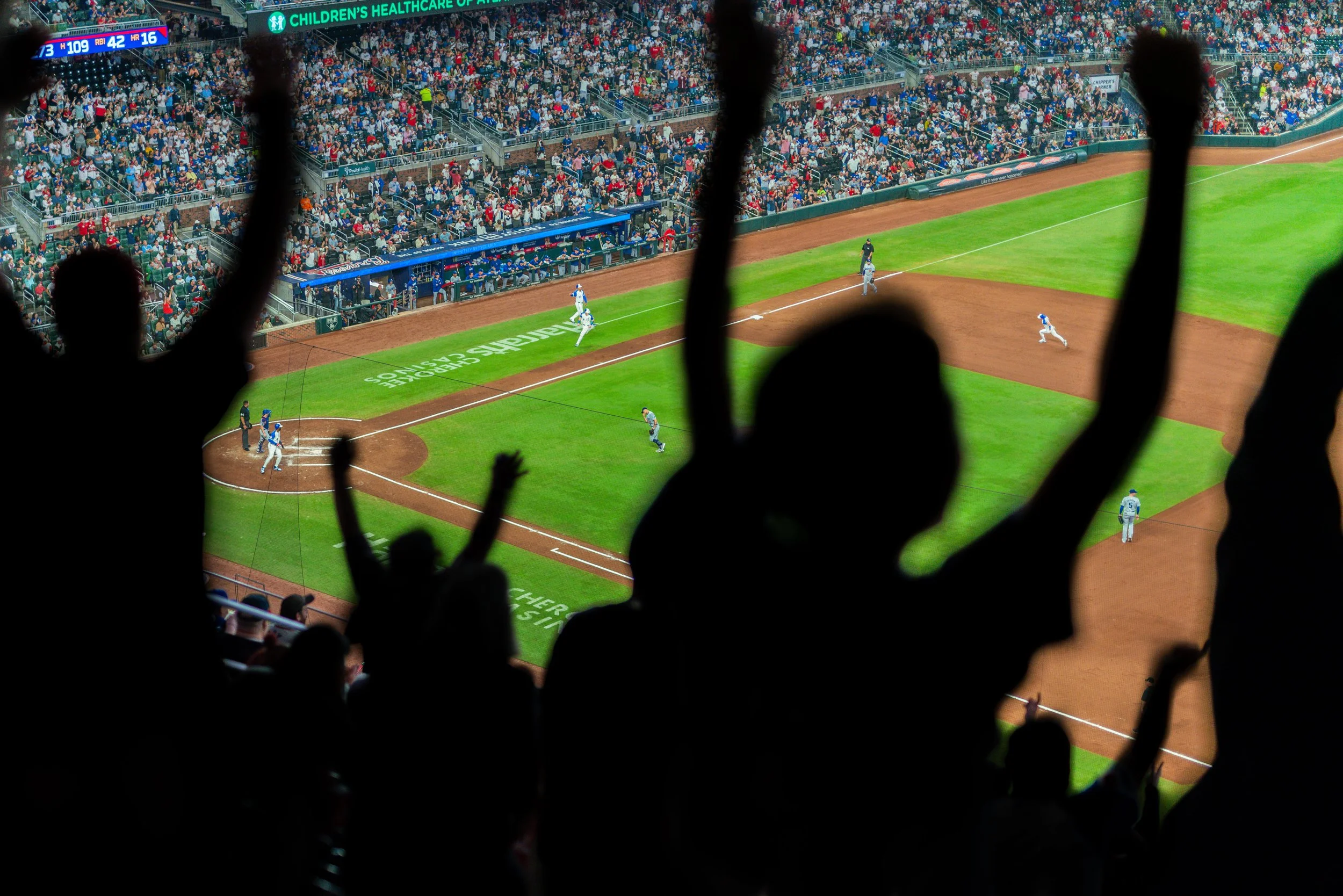 Crowd cheering at a baseball game, players running on the field with a full stadium in the background.