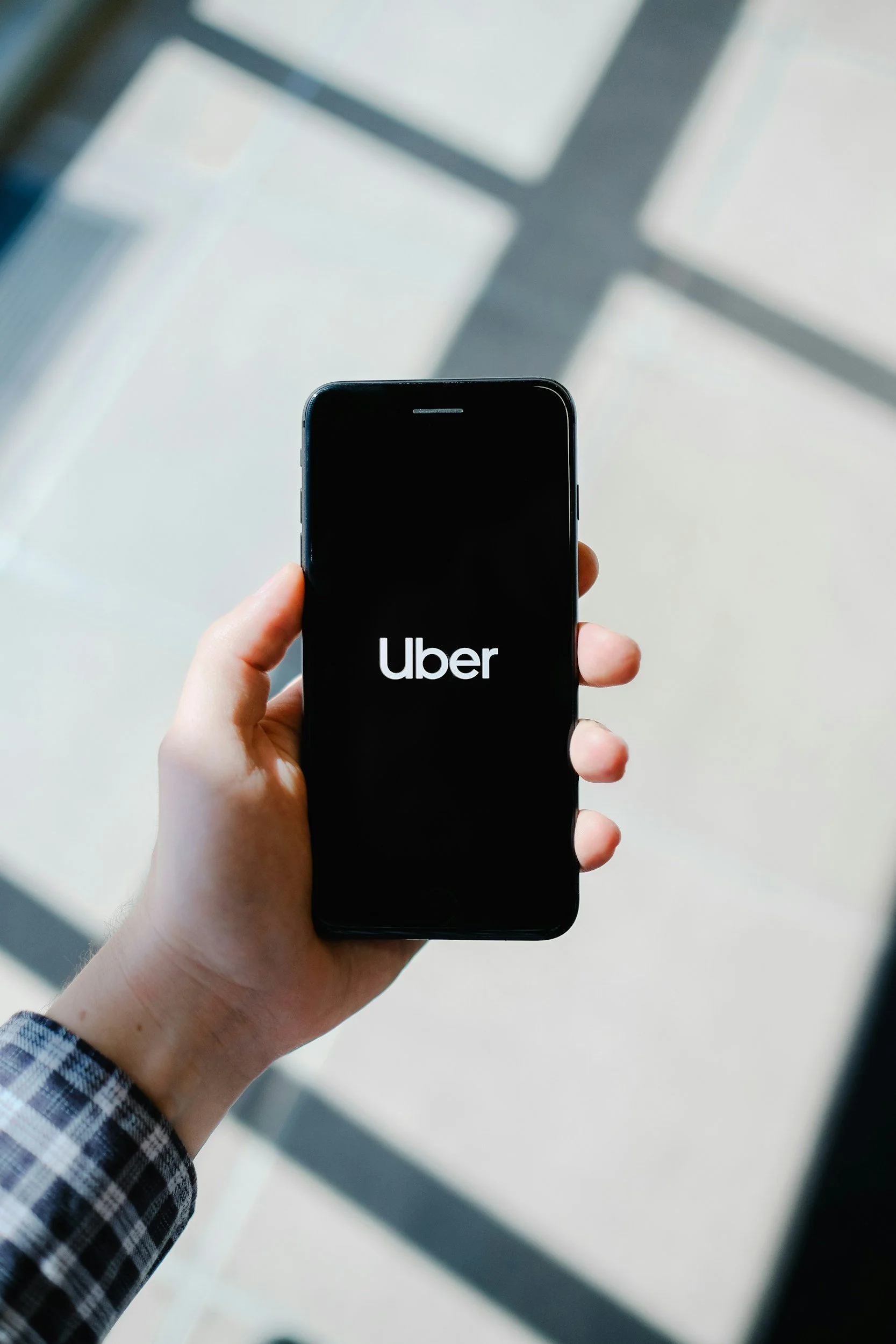 A person holding a smartphone with the Uber logo on the screen, with a shadow of a window cast on the floor in the background.